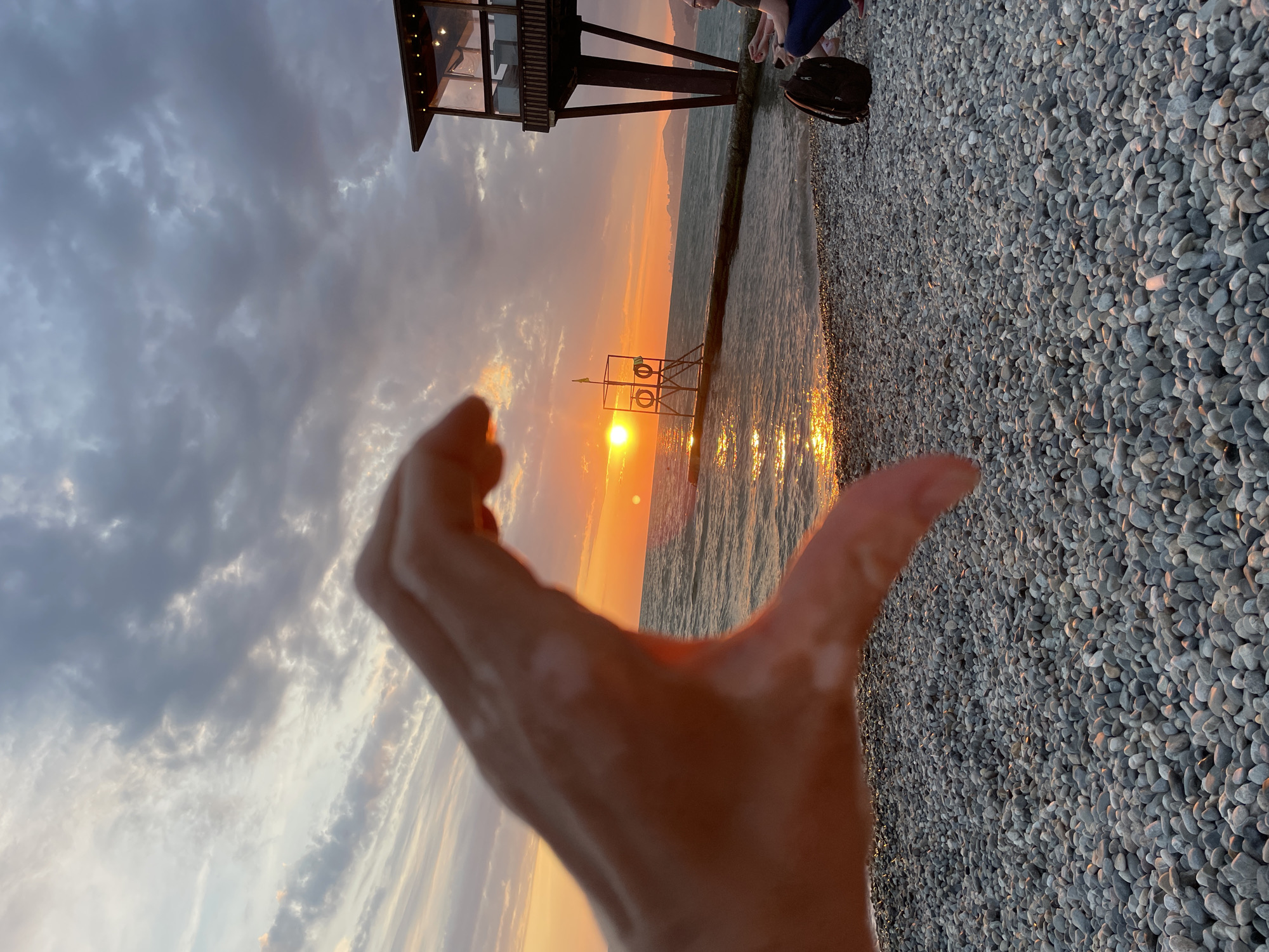 The image captures a sunset scene on a pebble beach, likely in Sochi, Russia, given the presence of a beachside structure resembling a cafe or restaurant on stilts. In the foreground, a person's hand is held up, forming a heart shape, with the setting sun visible through the opening. The sun, a bright orange orb, is positioned over the sea, casting a golden reflection on the water.
The beach is covered in smooth, grey pebbles. To the right of the frame, a dark backpack sits on the beach, and a person is partially visible, seated and looking out at the sea. In the middle ground, a pier extends into the water, supporting a lifeguard tower with two lifebuoys and a small flag. The sea is calm, with gentle waves lapping the shore. The sky is filled with dramatic clouds, tinged with orange and grey from the sunset. In the distance, a hazy coastline with mountains is visible. The overall atmosphere is serene and romantic, characterized by the warm hues of the sunset.