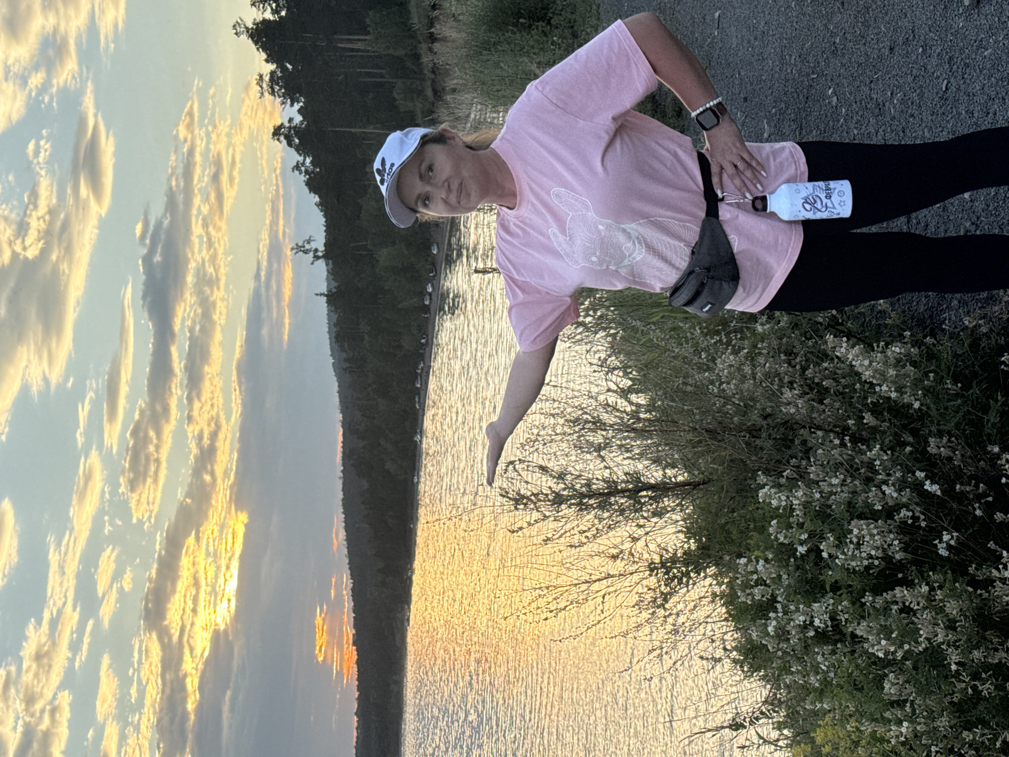 A woman stands in front of a body of water at sunset. She is wearing a pink t-shirt with a dog graphic, black leggings, a white baseball cap, and a grey fanny pack. A white water bottle with a blue design hangs from the fanny pack. Her left arm is extended, and her hand is open. The sun is setting over the water, casting a golden glow on the clouds and the water's surface. Trees line the distant shore. The scene appears to be a park or recreational area. This location is the Angara River embankment in the city of Bratsk, Russia.