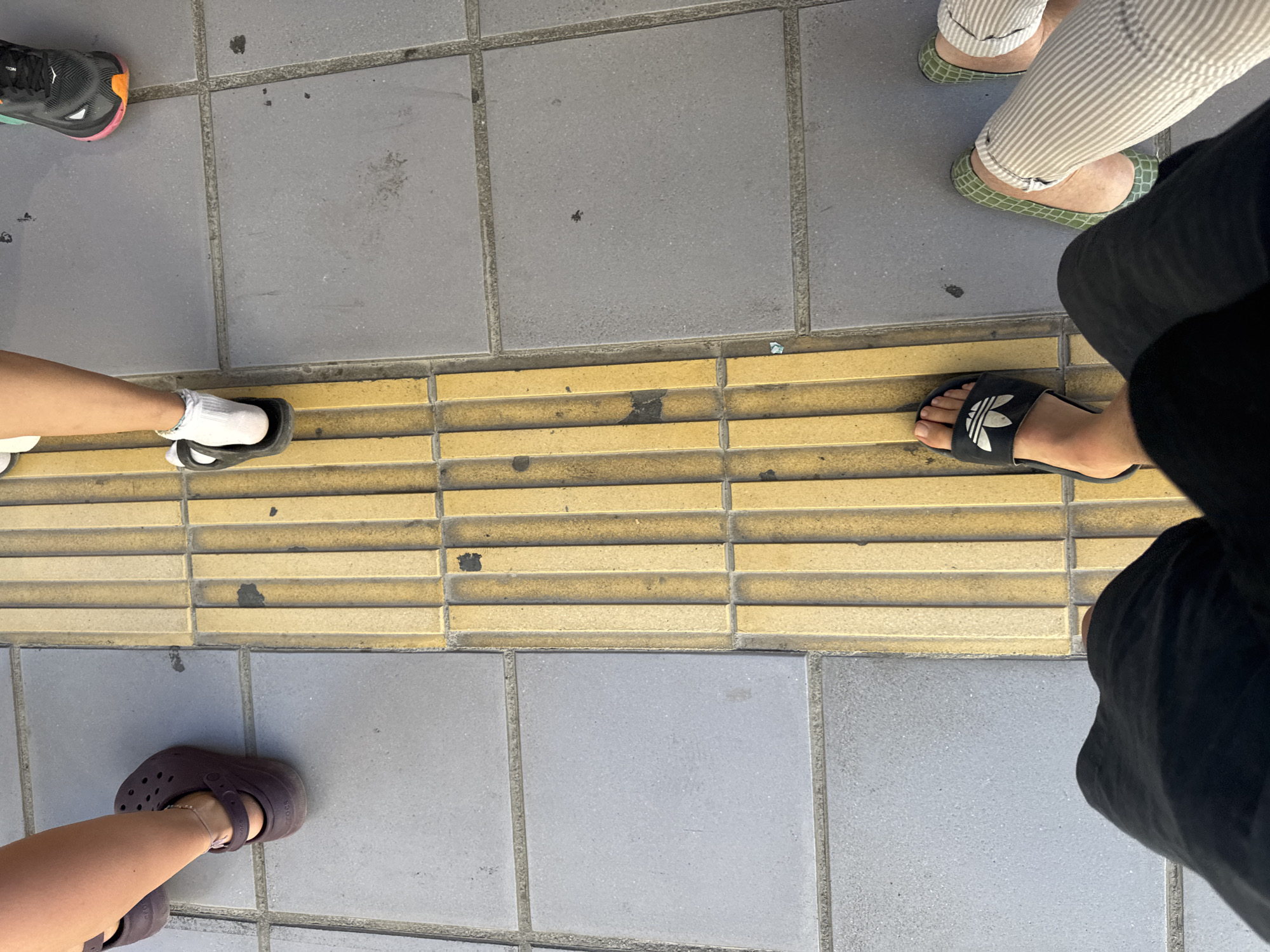 The image shows a close-up view of several people's feet and lower legs standing on a sidewalk in Chania, Greece. A yellow tactile paving strip, characteristic of pedestrian walkways and crossings in many cities, runs vertically through the center of the frame. The sidewalk is constructed of gray square tiles.
From left to right, the feet visible are:
- A person wearing dark brown Crocs.
- A person wearing black slide sandals with white socks.
- A person wearing black athletic shoes with a pink and orange accent on the sole.
- A person wearing black slide sandals with the Adidas logo.
- Two people wearing striped trousers, one with green patterned shoes and the other wearing white shoes.
The lighting suggests it is daytime, and the context of the tactile paving indicates an urban environment, likely a street or public square in Chania. The arrangement of feet suggests multiple individuals are standing or waiting, possibly interacting or in transit.