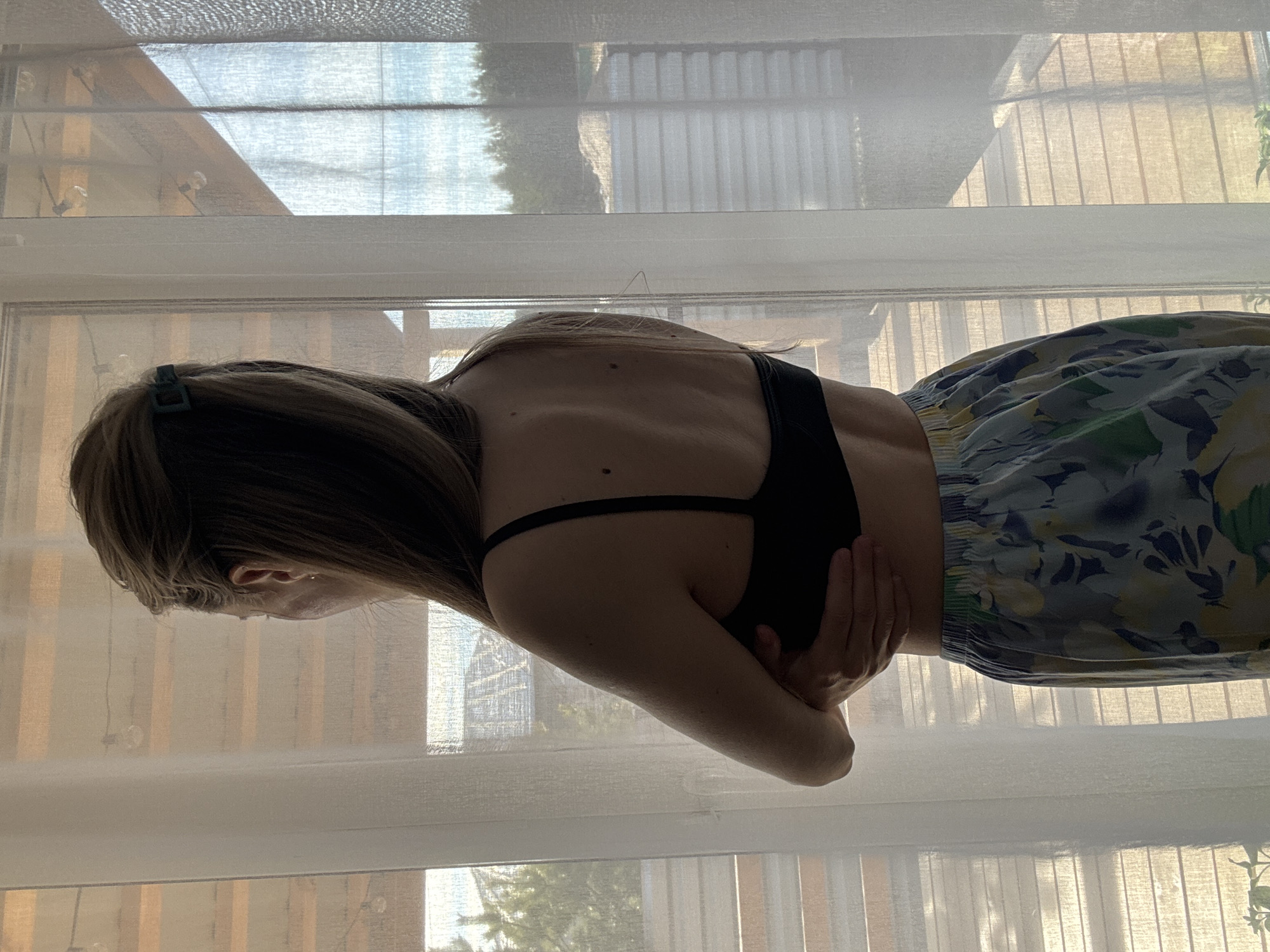 The image displays a person, viewed from the back, standing indoors by a large window. The individual, likely female with long brown hair, which is partially secured by a dark hair clip, wears a dark bra or fitted top and a flowing skirt or shorts adorned with a light-colored floral or abstract pattern featuring blue, yellow, and green hues. Their arms are crossed over their midsection, hands clasped around the opposite elbows, and their head is tilted downwards or away from the camera.
The person exhibits a lean physique, with visible shoulder blades and spinal definition on their back, along with several small moles. The background is dominated by a window covered with translucent, light-colored blinds or curtains, allowing diffused natural light to filter into the room. Through these semi-transparent coverings, the blurry outlines of trees or foliage, and potentially distant structures, can be discerned outside, indicating daytime. The natural light creates a soft silhouette effect on the person. No text is visible in the media file. The location context provided is Chaikovsky, Russia.