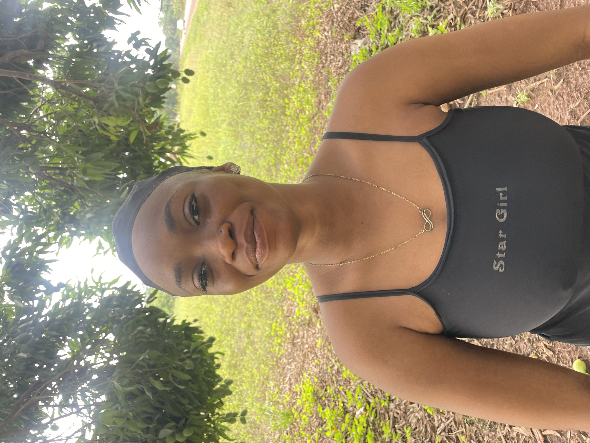 A young woman is positioned outdoors in what appears to be a park or natural setting in Ogpe, Nigeria. She is wearing a black tank top with the words "Star Girl" printed on it and a gold necklace with an infinity symbol pendant. She also has a black head covering and an earring. The background is filled with green grass and foliage, including a tree with leaves and some immature fruits. The ground shows a mix of grass and brown dirt. The overall lighting suggests it is daytime.