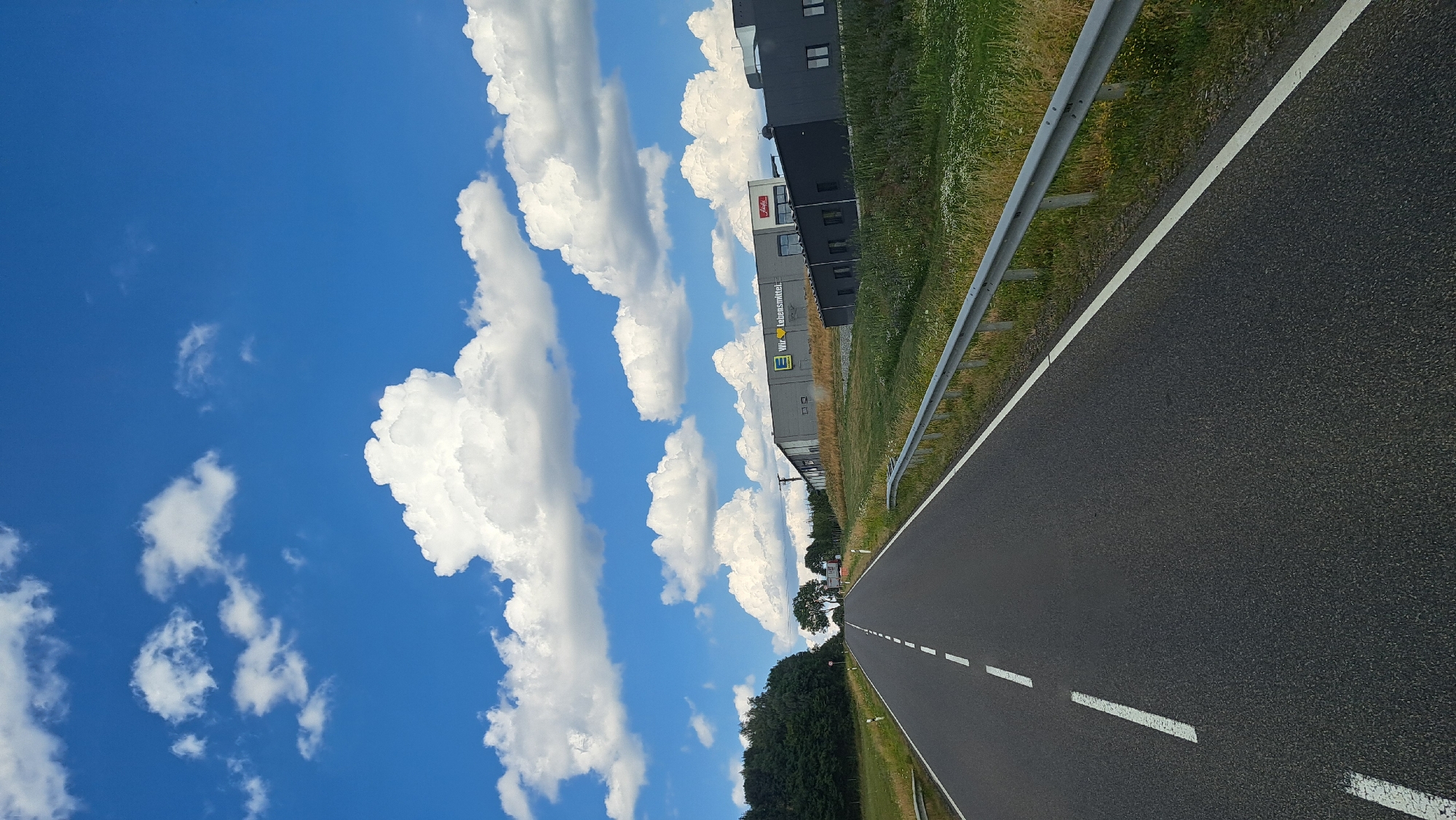 The image depicts a view from a moving vehicle on a two-lane asphalt road in Höhn, Germany. The road is marked with white dashed lines on the left lane and a solid white line on the right edge, indicating a clear path forward. A metal guardrail runs along the right side of the road, separating it from a grassy verge dotted with wildflowers.
In the background, to the right of the road, are several modern industrial buildings. One building prominently displays the yellow "E" logo of an Edeka supermarket chain, with the text "Wir leben Lebensmittel" (We live groceries) written beside it. Another building to its right is dark grey and appears to be a warehouse or commercial property.
The landscape to the left of the road is characterized by a dense line of green trees, forming a forest edge. Beyond the trees, the sky is a vibrant blue, filled with large, fluffy white cumulus clouds. The overall scene suggests a sunny day in a rural or semi-industrial area outside of the main town center of Höhn. There are no people or animals visible in the frame.