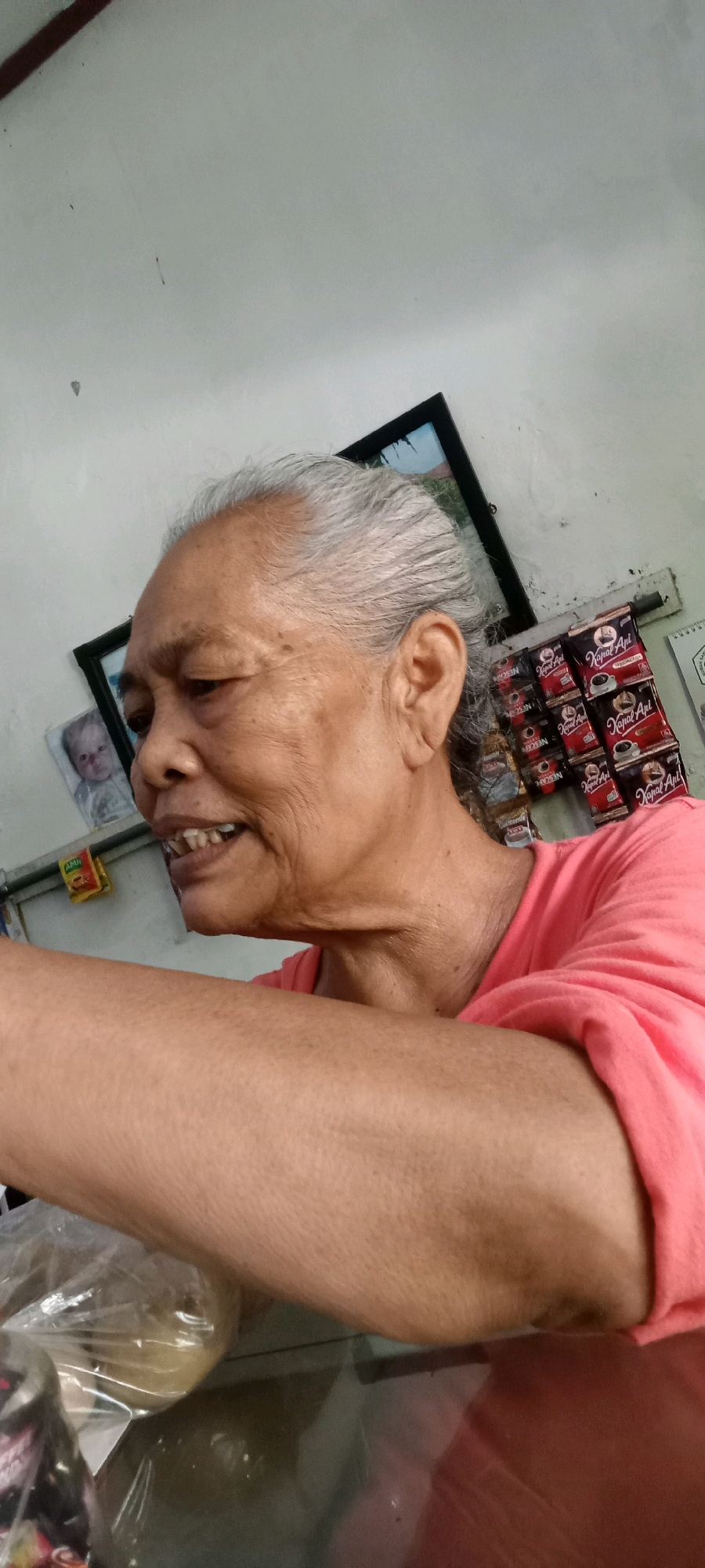 The image shows a close-up, profile view of an elderly Indonesian woman with gray hair, wrinkles, and missing teeth, wearing a coral pink shirt. She is in what appears to be a domestic setting, possibly Jaten, Indonesia, with a white, textured wall in the background. Behind her, a framed picture of a baby is visible, along with several packets of instant coffee labeled "Kapal Api" and "Nescafe." The woman's right arm and shoulder are in the foreground, and a plastic-wrapped object is partially visible on a glass surface. The lighting is somewhat dim, with a slight glare on the glass surface.