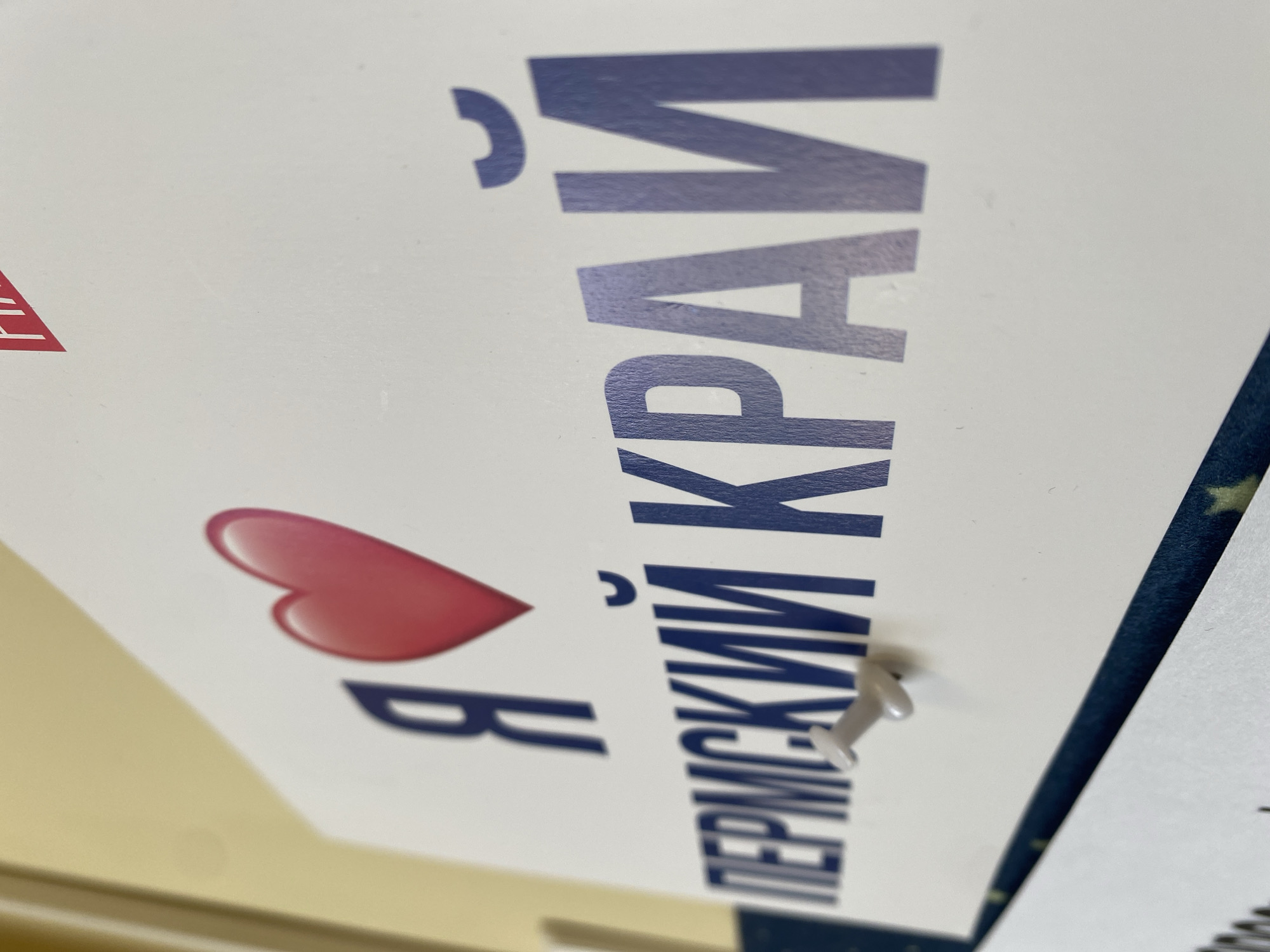 A close-up shot shows a poster angled vertically. The poster displays the phrase "Я ❤️ ПЕРМСКИЙ КРАЙ" (I love Perm Krai) written in Russian. A red heart graphic replaces the word "love." The text is dark blue. A pushpin is visible, securing the poster in place. The context suggests this might be located in Chaikovsky, Russia, given the reference to Perm Krai, a region within Russia where Chaikovsky is situated.