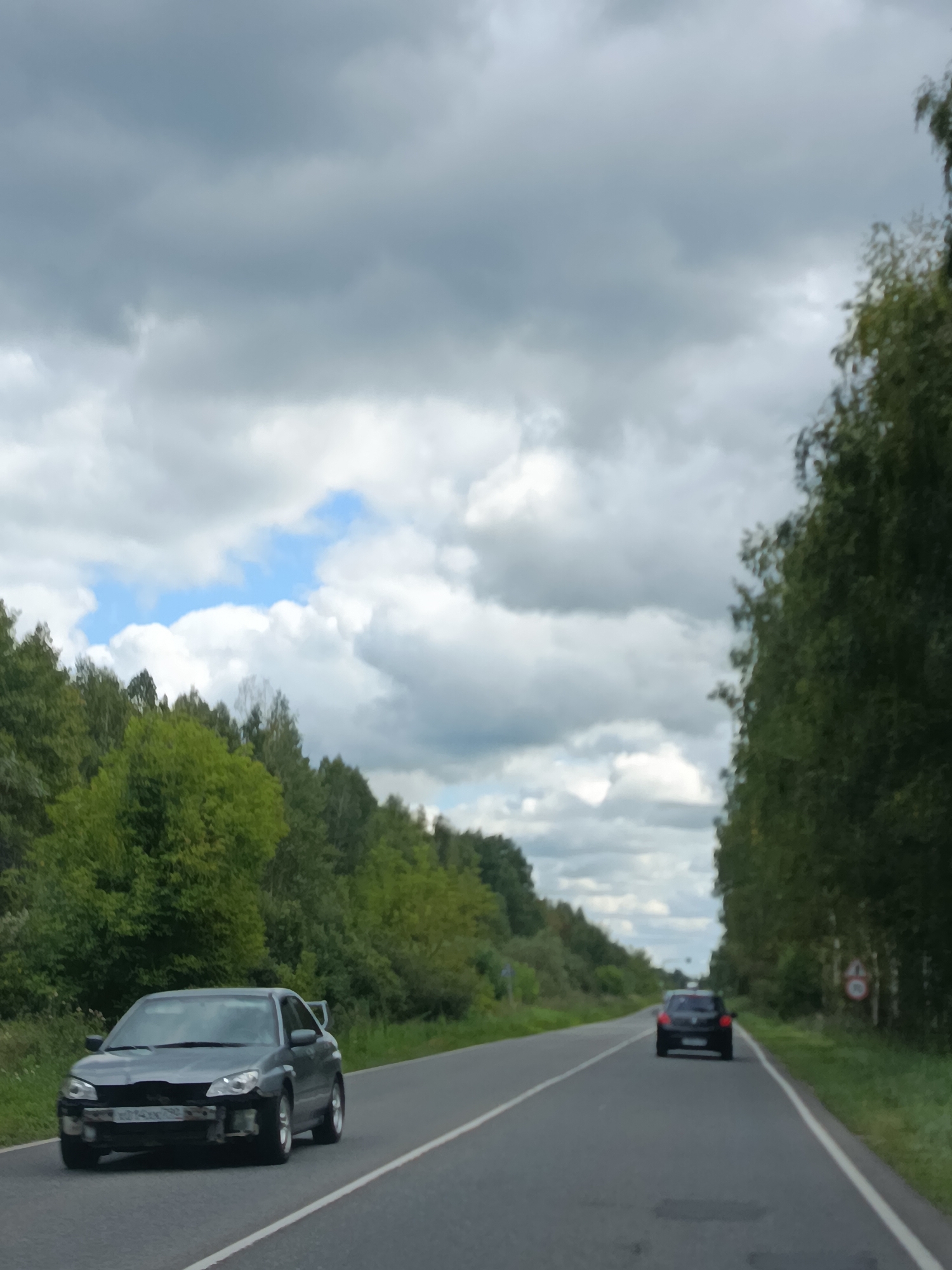 In an outdoor, daytime scene on a two-lane asphalt road, a dark gray sedan with damage to its front bumper and grille is driving in the left lane, facing the camera. The car is positioned in the foreground and to the left of the frame. The road stretches into the distance, flanked by dense green trees on both sides. In the background, a black hatchback is driving away from the camera in the right lane. The sky above is filled with a mix of gray and white clouds, with patches of blue sky visible. A speed limit sign is visible on the right side of the road, behind the black hatchback. The setting appears to be a rural or semi-rural area, possibly a highway or main road surrounded by woodland.