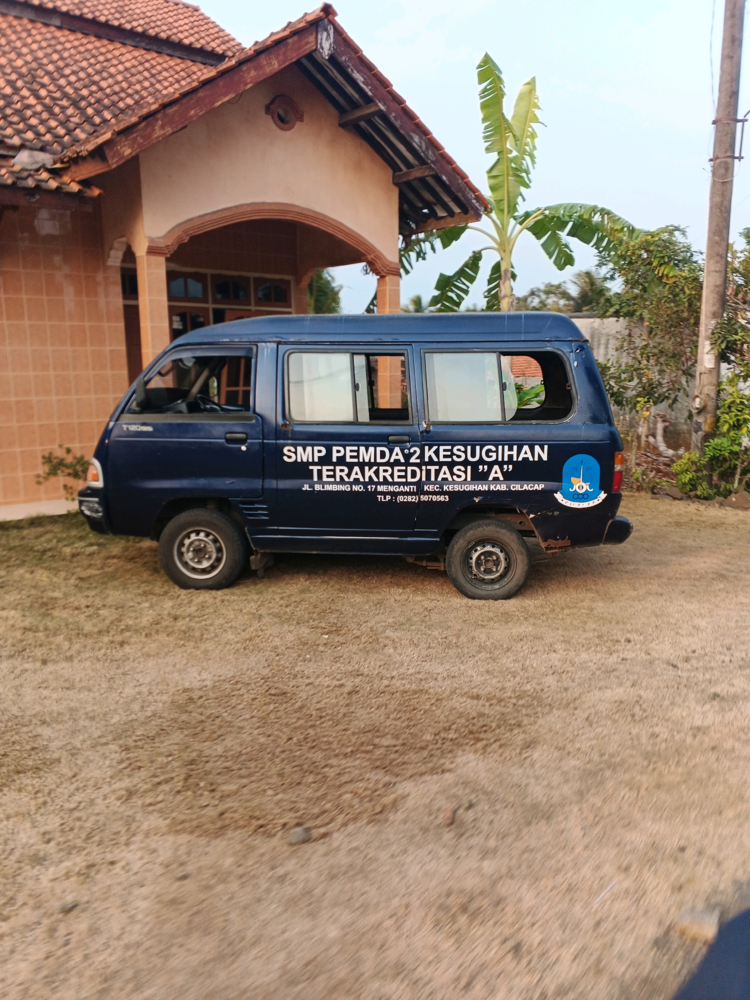 A dark blue, four-door van is parked on a dirt and grass lot in front of a light brown, tiled house with a reddish-brown tiled roof. The van has white lettering on its side that reads "SMP PEMDA 2 KESUGIHAN TERAKREDITASI "A"" and below that "JL. BLIMBING NO. 17 MENGANTI . KEC. KESUGIHAN KAB. CILACAP TLP: (0282) 5070563". A blue and white logo is also visible on the rear passenger side of the van. To the left of the van is a porch with a brown arched entryway leading into the house. Behind the van, a tall plant with large green leaves, likely a banana tree, grows. A concrete wall and trees are visible in the background, as well as a utility pole on the right side of the image. The sky is overcast and pale.