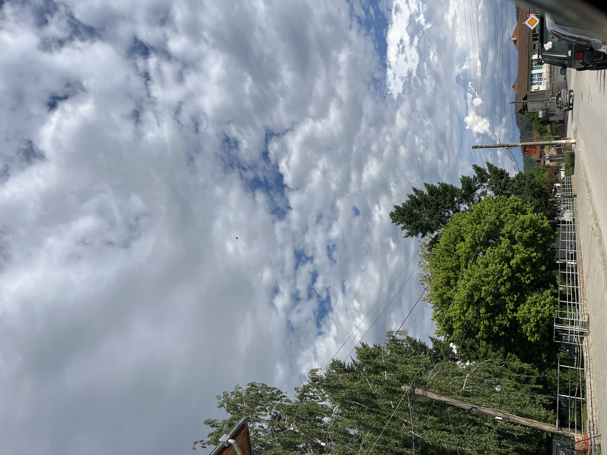 The image shows a view of the sky with scattered cumulus clouds, taken from a street in Kornitsa, Bulgaria. A portion of a building with a brown roof is visible on the right edge of the frame, adjacent to the sky. There is a road and a car parked on the road. Trees are present, and utility poles with wires run across the sky. A section of a white metal fence runs along the lower right of the frame.