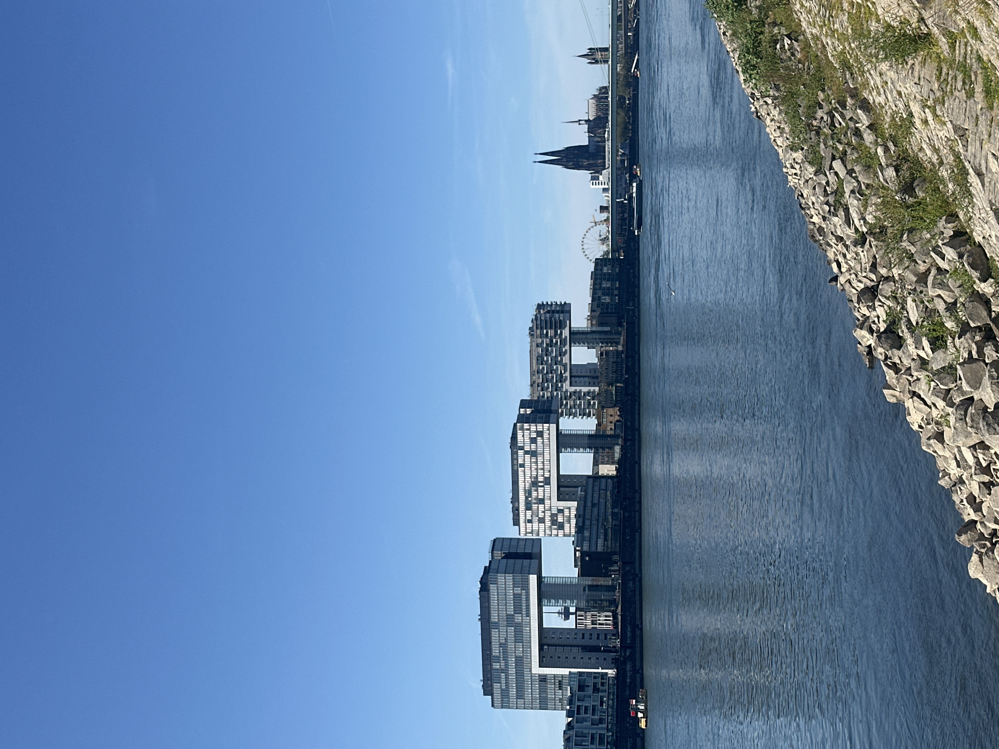 This is a photograph of the Rhine River in Cologne, Germany, on a clear day. The foreground shows a rocky embankment leading down to the blue water. In the middle ground, a large river spans the width of the frame. On the far bank, a prominent cityscape unfolds, dominated by modern architecture, including the Kranhäuser, a cluster of three distinctive crane-shaped buildings. Further to the right, the iconic Cologne Cathedral with its twin spires stands tall, along with other historical and modern structures. A Ferris wheel is also visible in the distance. Several boats are navigating the river. The sky is a vast expanse of clear blue.