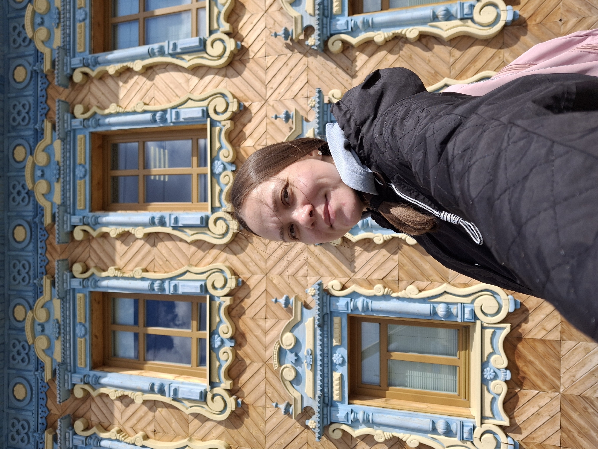 A fair-skinned woman with light brown hair, tied back, is positioned in front of a wooden building in Sarapul, Russia. The building has intricately carved window frames painted in blue and beige. The wooden planks forming the facade are arranged in a herringbone pattern. The woman is wearing a black quilted jacket with a light blue hood visible, and a light pink backpack.