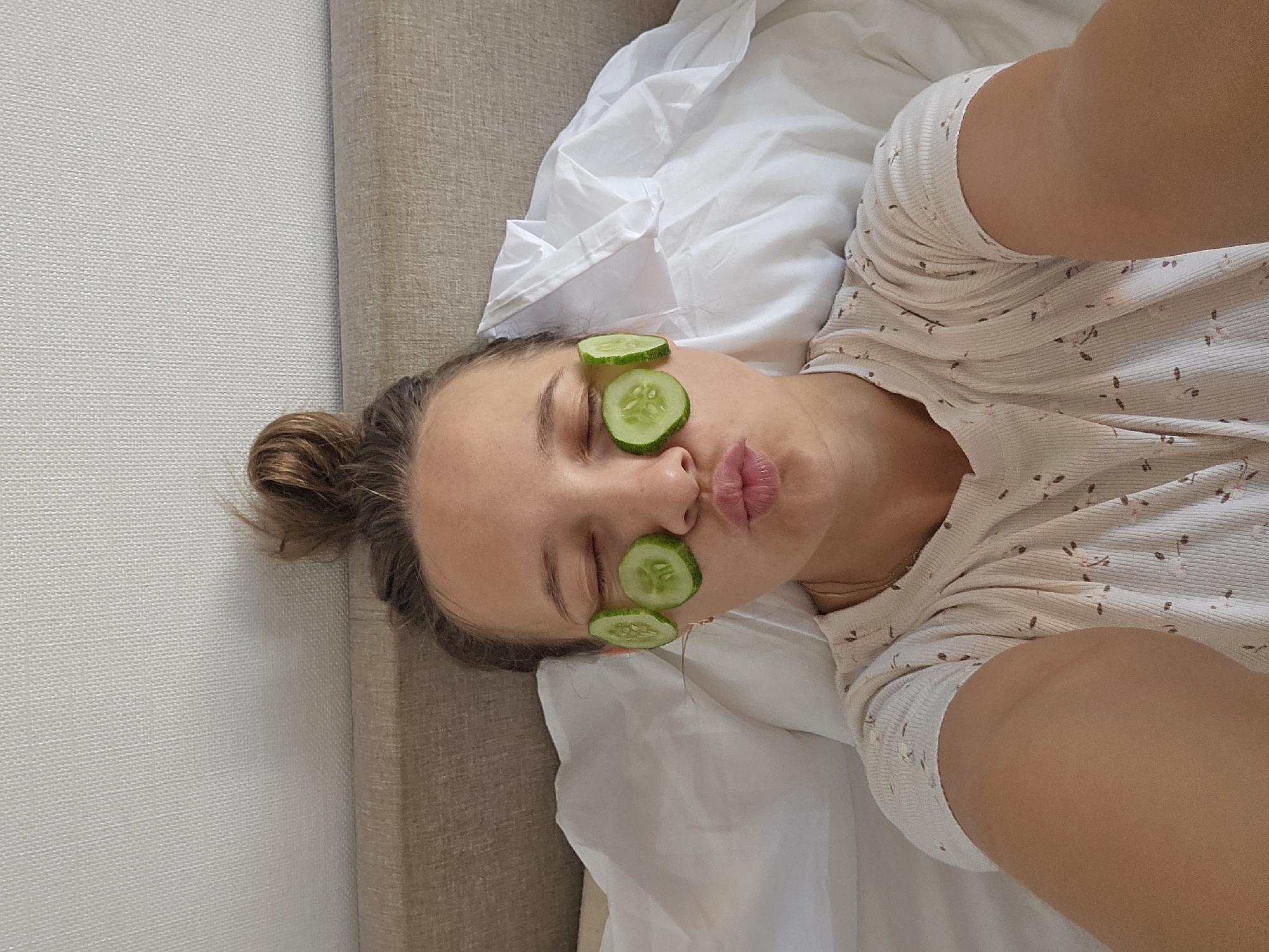A young woman with her hair in a bun is lying in bed, with cucumber slices on her closed eyes. She is wearing a light-colored t-shirt with a floral pattern. The room is softly lit, and the background consists of a white duvet and a textured headboard.