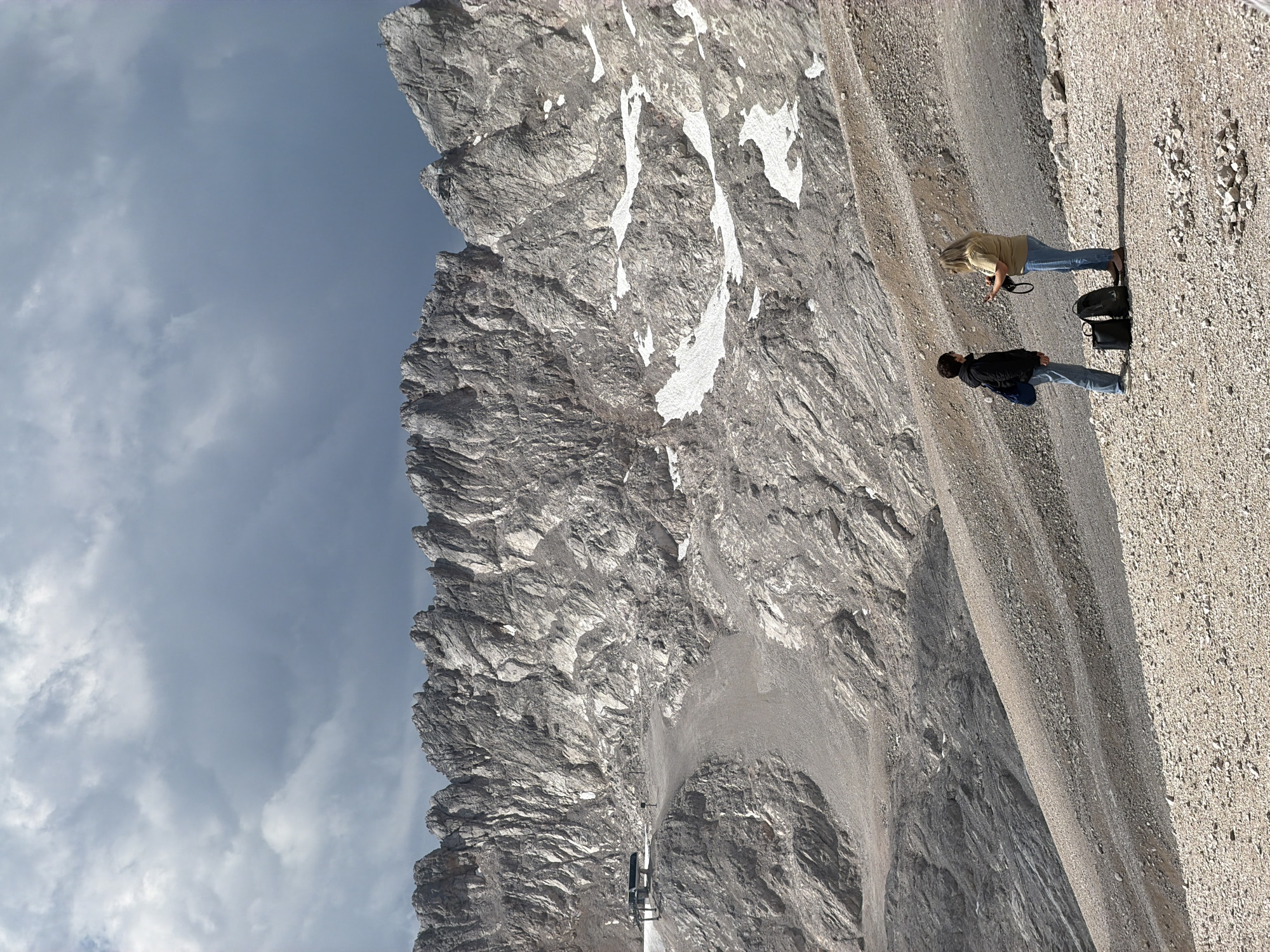 The image depicts a panoramic view of a mountainous landscape, likely in the vicinity of Garmisch-Partenkirchen, Germany, characterized by rugged, rocky peaks and scattered patches of snow. The sky is overcast with dark, dramatic clouds. In the foreground, two individuals, one wearing a dark jacket and the other a light-colored shirt, are standing on a gravelly slope. They are positioned facing each other, engaged in conversation. Beside them, two backpacks and a handbag rest on the ground. Further up the slope, a cable car station is visible, serving the mountainous terrain. The environment is arid and rocky, with sparse vegetation, typical of high-altitude mountainous regions.