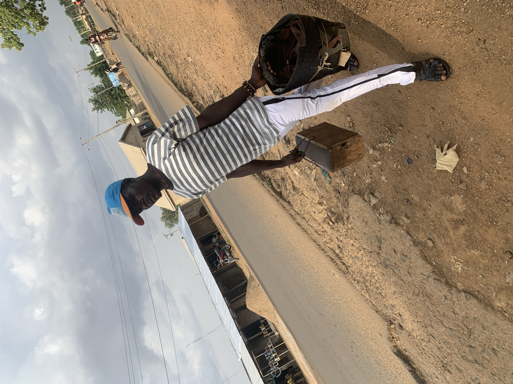 A man stands on the shoulder of a road in Maigizo, Nigeria. He wears a blue baseball cap, striped shirt, and white pants. He holds a wooden box in his right hand and a dark bag in his left. He is wearing sandals. The road has a building adjacent to it. Power lines stretch overhead. The road is lined with brown dirt. The sky is cloudy.