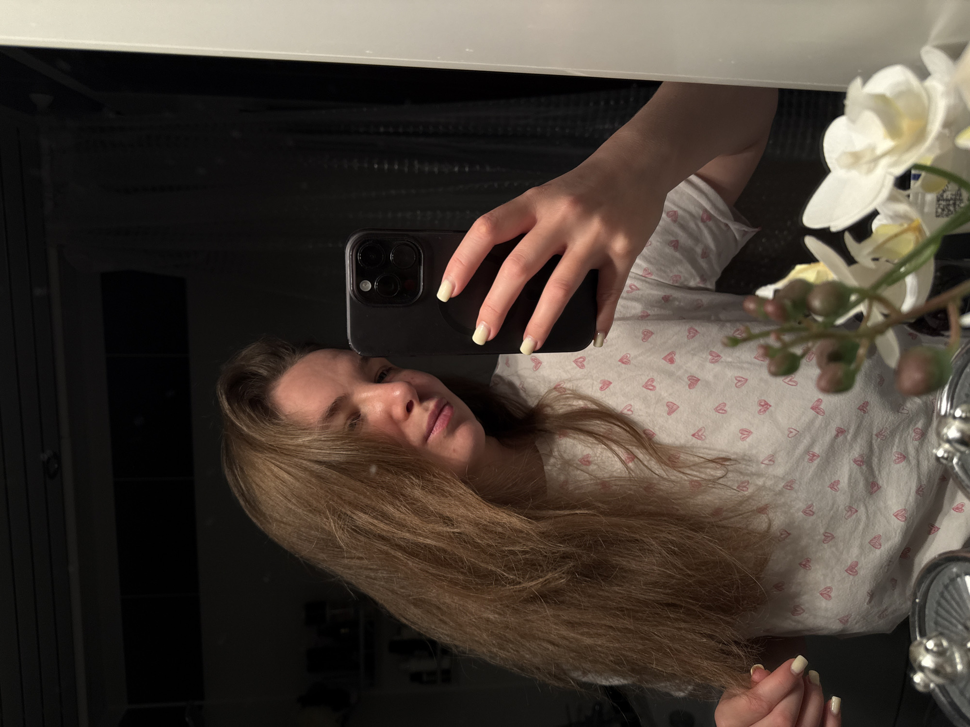 A woman with long, light brown hair is taking a selfie in a mirror. She is wearing a white t-shirt with small pink heart patterns. Her nails are painted a pale yellow. She is holding a black smartphone. To the right of the frame is a white orchid in a vase. The reflection suggests the location is indoors, possibly a bathroom or bedroom. The environment appears to be domestic and well-lit. There is no verifiable indication of Vladimir, Russia.