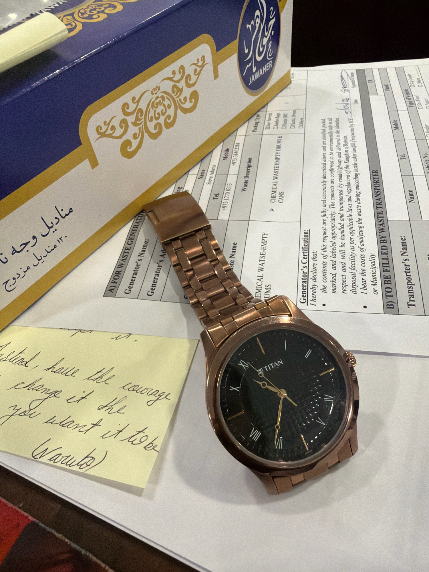 An overhead shot captures a desk surface with several items, including a watch, a yellow sticky note, a blue and gold patterned box, and a document. The watch, a Titan, is made of rose gold with a black face and is positioned prominently in the foreground. A yellow sticky note with handwritten cursive text is placed to the left of the watch. The text appears to be a motivational quote. To the left of the sticky note and watch, a portion of a blue box with intricate gold Arabic calligraphy and floral designs is visible. The box has the word "JAWAHER" printed on it in a stylized font. A white document with printed forms and handwritten entries is partially visible beneath the watch and box, suggesting it is a work or logistical document. The overall scene suggests a casual workspace or office environment.