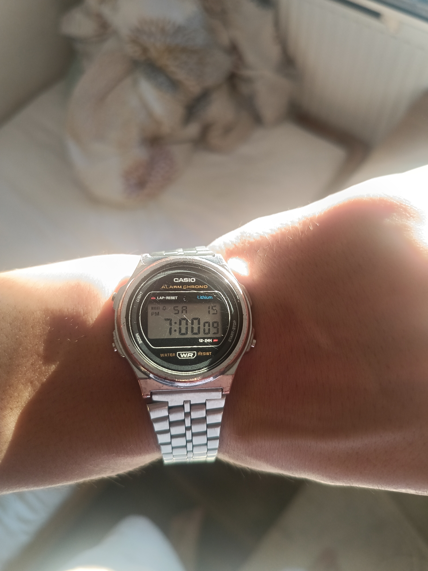 The image shows a close-up of a person's wrist wearing a silver Casio digital watch. The watch face displays the time as 7:00:09, the date as Saturday, the 5th, and the month as April (indicated by "SA" and "15"). The watch also shows "PM" and "12-24H" indicators. The brand name "CASIO", "ALARM CHRONO", "Lithium", "LAP-RESET", "WATER WR RESIST" are visible on the watch face. The background is out of focus, showing a blurred white and beige patterned surface, possibly a blanket or fabric. The lighting suggests the photo was taken in natural sunlight, casting a strong glare on the watch and the person's skin. The location of Grodzisk Mazowiecki, Poland is not visually identifiable from the image content.