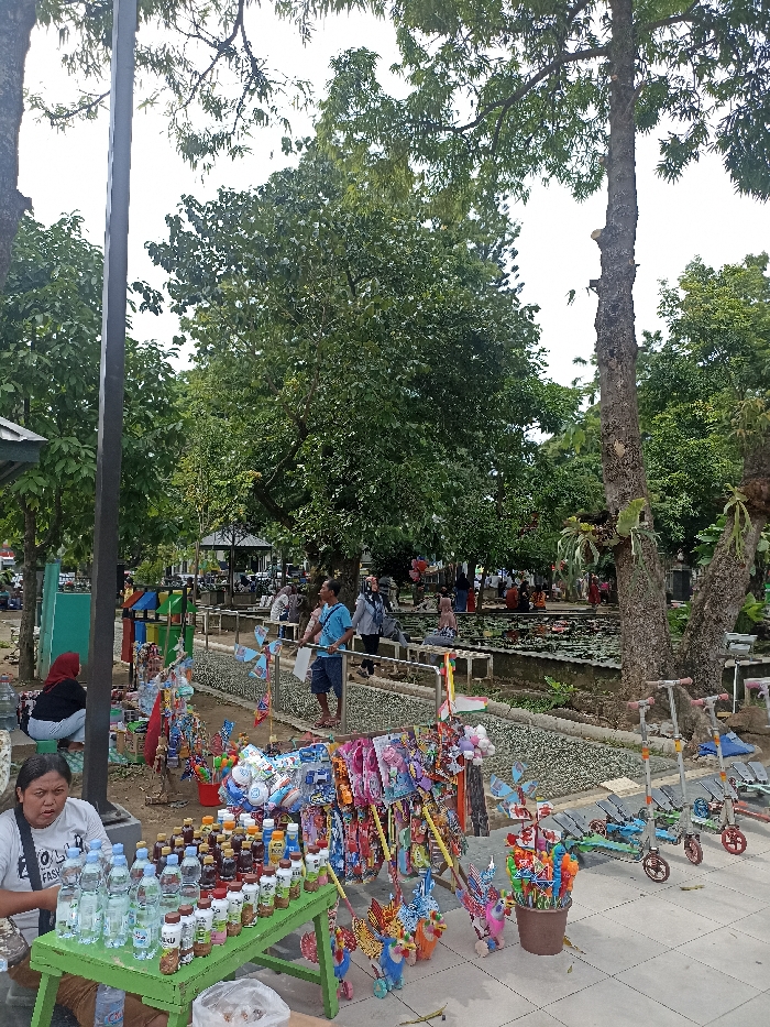 This image depicts an active public park scene in Tulungagung, Indonesia, during daytime under an overcast or partly cloudy sky. The setting is lush with numerous large trees providing shade, and a pond filled with lily pads is visible in the mid-ground.
In the foreground, a vendor stall is set up on a paved surface. A woman wearing a white t-shirt with the word "FASHION" visible is seated behind a green table, offering various bottled drinks, including water and other beverages. To her right, a vibrant display of children's toys such as pinwheels, colorful character figures, and other trinkets are arranged for sale or rent. Three child-sized kick scooters are also parked nearby. Another woman in a red hijab is seen crouching behind the vendor's stall. Colorful, segmented waste bins are positioned behind them.
In the mid-ground, a pathway with a textured surface, possibly designed for reflexology, runs alongside the pond. Several people, mostly adults, are seen strolling, standing, and observing the surroundings along this path. Some are looking towards the pond, while others appear to be interacting. Further in the background, more people are gathered near additional park structures, suggesting various recreational activities. The overall scene conveys a relaxed and communal atmosphere in an urban green space.