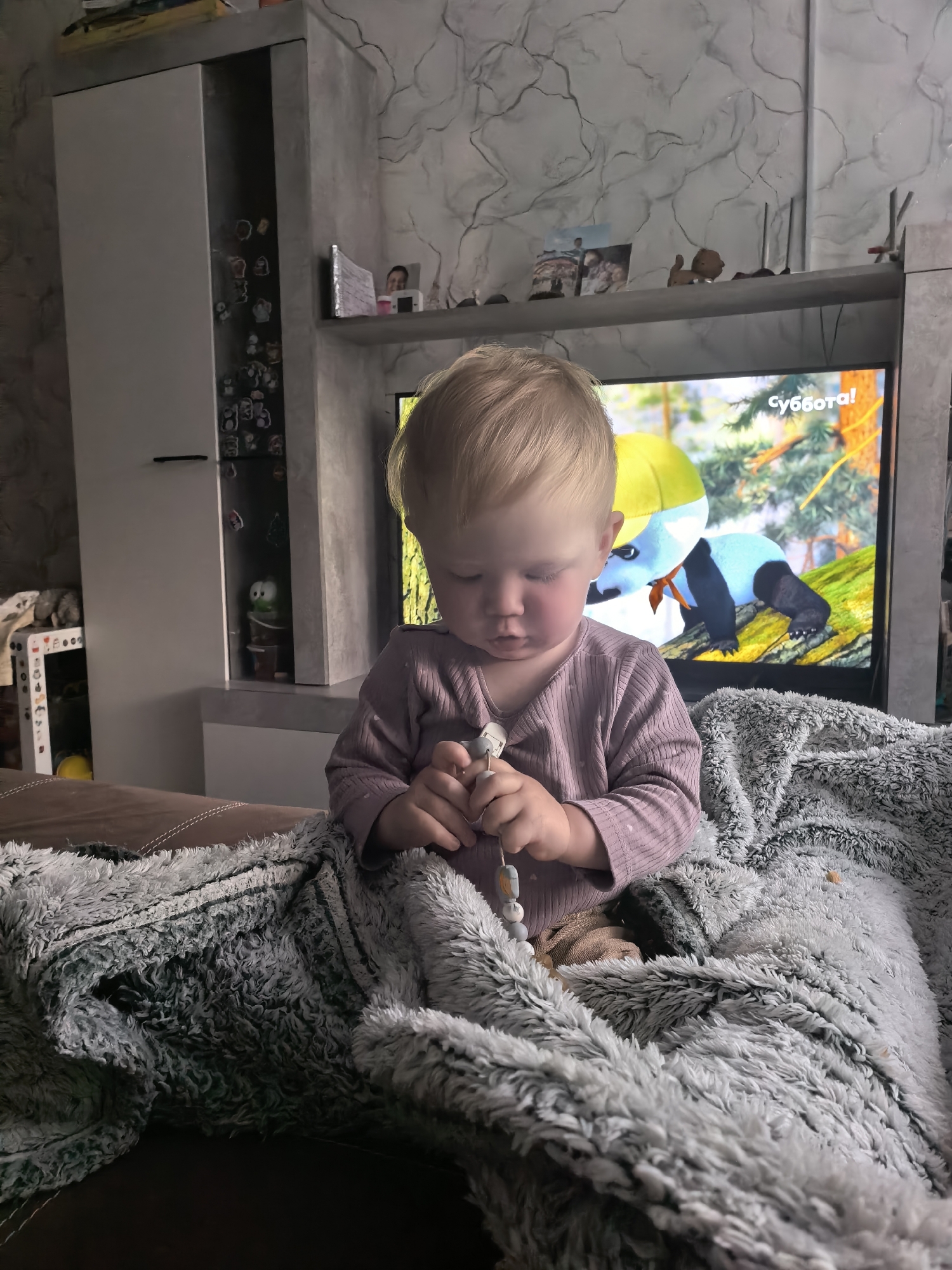 A young child with blonde hair is sitting on a couch, looking down at a toy. The child is wearing a light purple long-sleeved shirt and is partially covered by a gray, fluffy blanket. A television is on in the background, showing an animated panda wearing a yellow hat. To the left of the television is a white cabinet with glass doors, displaying various small items. The wall behind the television has a textured, marbled pattern. The scene appears to be indoors, likely in a living room. There is no information to suggest the location is in the city of Sterlitamak, Russia.