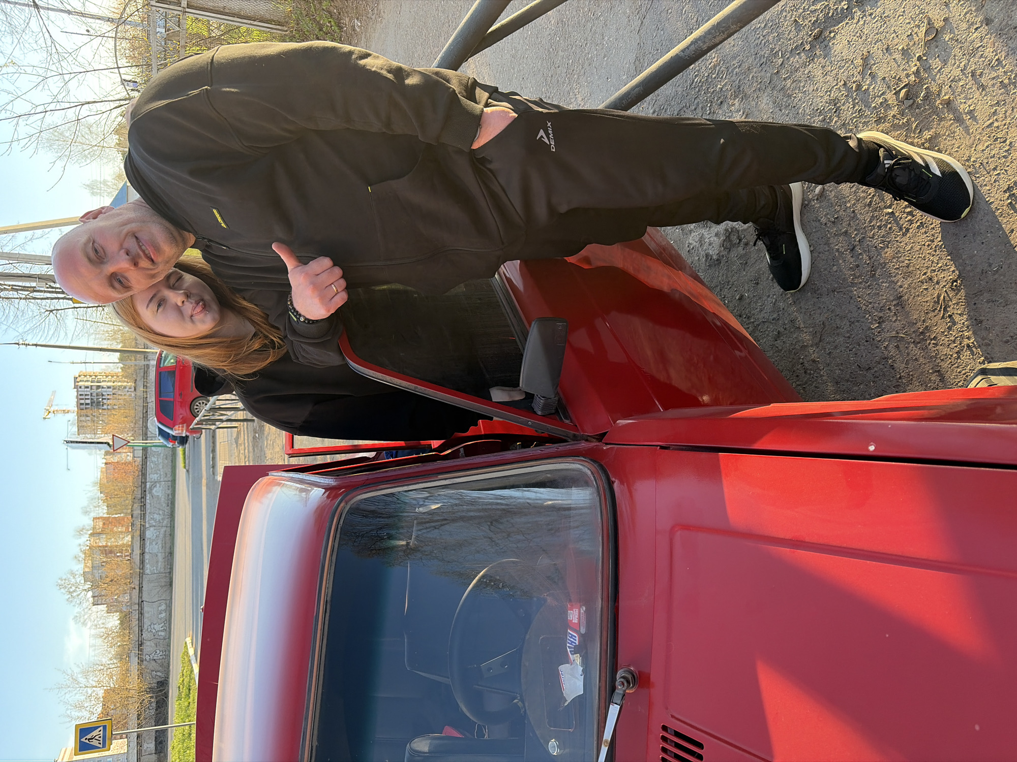 A middle-aged man and a younger woman are posing next to a red Lada car in Chaikovsky, Russia. The man is bald and wearing a black tracksuit with the logo "DEMIX" visible on the pants. He has one hand in his pocket and is giving a thumbs-up with the other. The woman has reddish-blonde hair and is sticking her tongue out slightly. The car's hood is open. In the background, there is a construction site, a street with a pedestrian crossing sign, and bare trees. A red car can also be seen parked in the background.