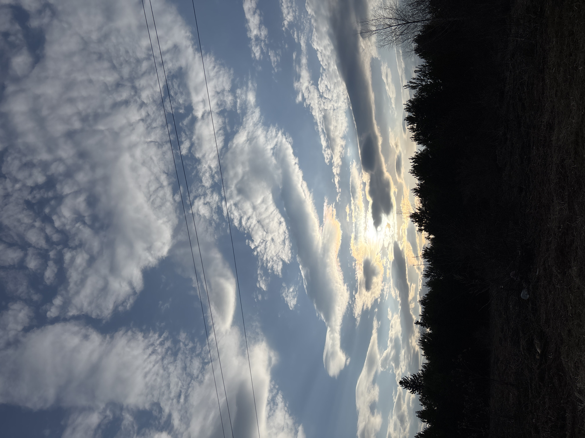 The image depicts a view of the sky with clouds, power lines, and silhouetted trees. The sky is blue with a mix of small, puffy white clouds and longer, more stretched-out gray clouds. Sunlight is visible through the clouds near the horizon. Power lines stretch across the left side of the frame. The right side shows a dark silhouette of trees against the bright sky. The location cannot be verified as Фоки, Russia, without more context or details within the image.
