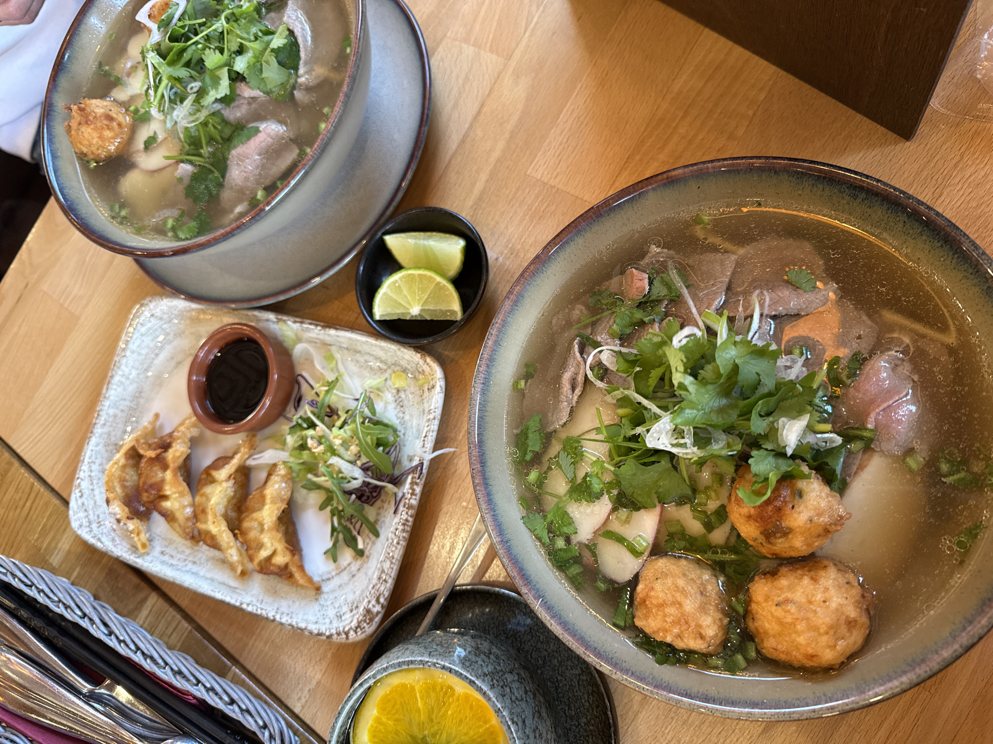 A top-down shot captures a lunch scene on a wooden table, likely in Senftenberg - Zły Komorow, Germany. Two bowls of soup, both containing meat, noodles, and garnished with fresh herbs, occupy the upper part of the frame. A rectangular dish with four fried dumplings arranged alongside a small bowl of dark sauce and a handful of greens is positioned near the lower left. A small black bowl holding two lime wedges rests between the dumplings and one of the soup bowls. A cup containing a slice of orange is visible at the bottom center. A basket with silverware peeks into the frame at the lower left. The lighting is bright and even, highlighting the textures of the food and the table's wood grain.