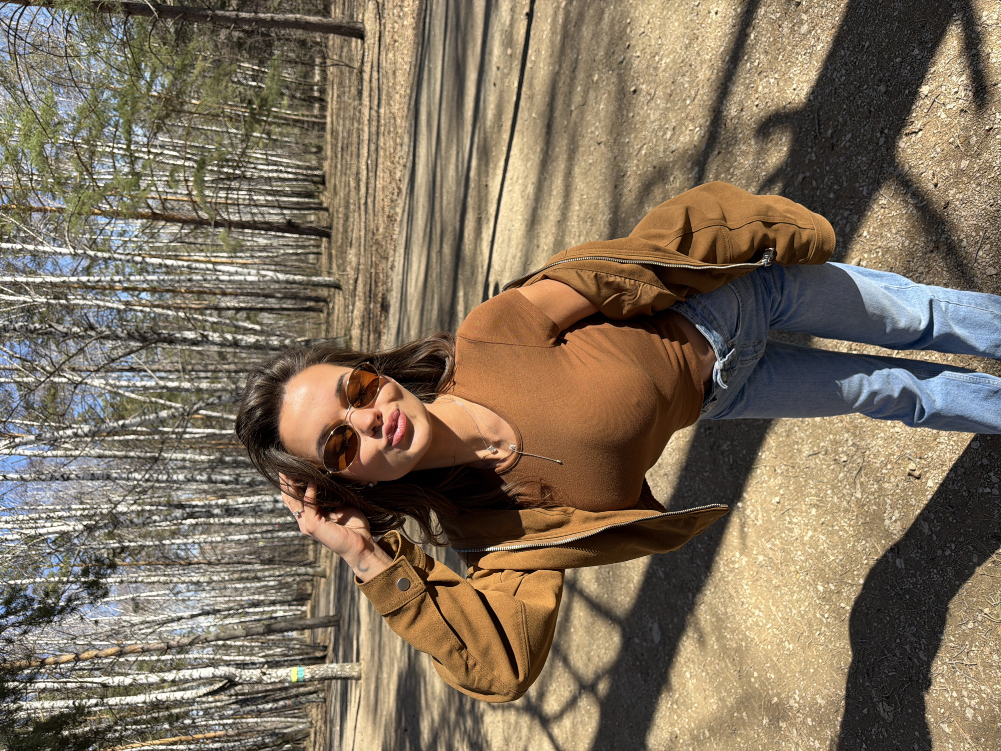 A young woman is captured in a full-body shot, posing on an unpaved path in a forest or park setting, likely in Krasnoyarsk, Russia. The image is taken on a bright, sunny day, as evidenced by the distinct, long shadows cast across the ground.
The woman has long, dark hair and is wearing amber-tinted, round sunglasses. She is dressed in a fitted, long-sleeved brown top, a matching brown zip-up jacket, and light blue jeans. A delicate silver necklace with a small pendant is visible around her neck. She looks towards the camera with a soft expression, her lips slightly parted in a subtle pout or smile. Her left hand is casually raised, touching the side of her head or sunglasses. A small, dark tattoo is visible on her left forearm/wrist.
The background features a dense concentration of trees. Predominantly, numerous birch trees with their characteristic white bark and slender trunks line the area, many appearing to be bare of leaves, suggesting either early spring or late autumn. Some coniferous trees with green needles are also visible among them. The ground is a mix of dirt, small stones, and dry foliage. The overall scene conveys a relaxed outdoor moment in a natural, wooded environment. No visible text is present in the image.