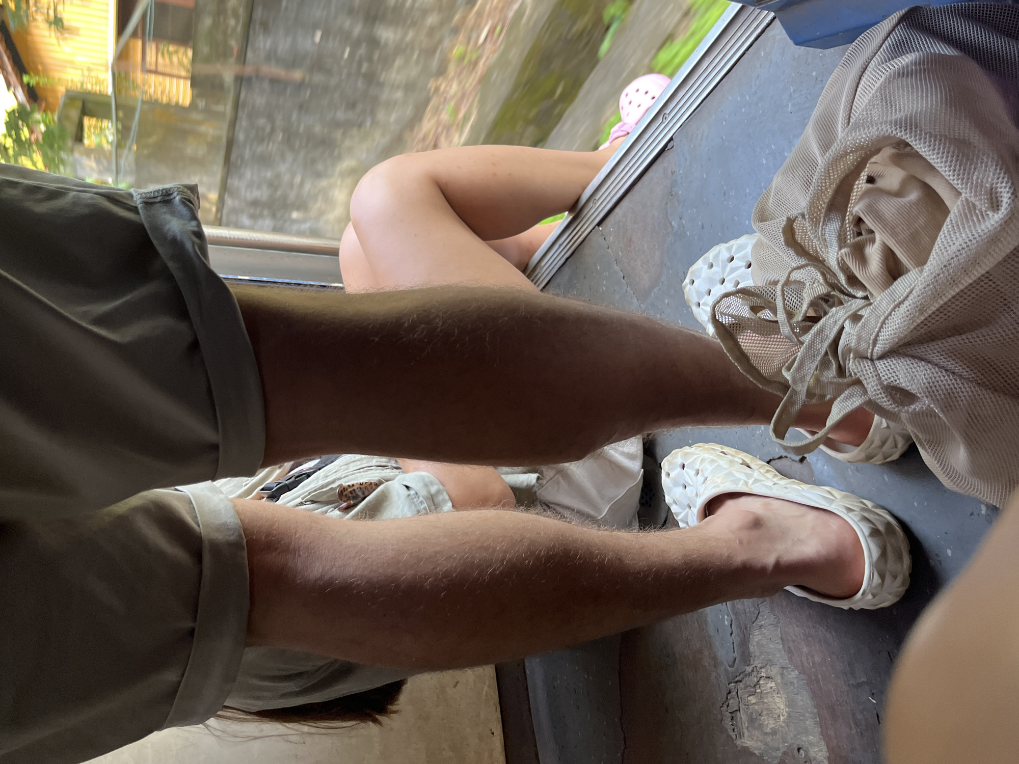 The image shows the lower legs and feet of two individuals inside a vehicle, possibly a train or bus, in Ambalangoda, Sri Lanka. The person in the foreground on the left has hairy legs and is wearing white, textured clogs. Their shorts are rolled up. To the right, the lower leg and knee of another person are visible, wearing pink clogs and sitting with their legs extended towards an open window. Outside the window, a blurred background of greenery and buildings suggests an urban or semi-urban environment. A mesh bag with clothing is also visible in the foreground.