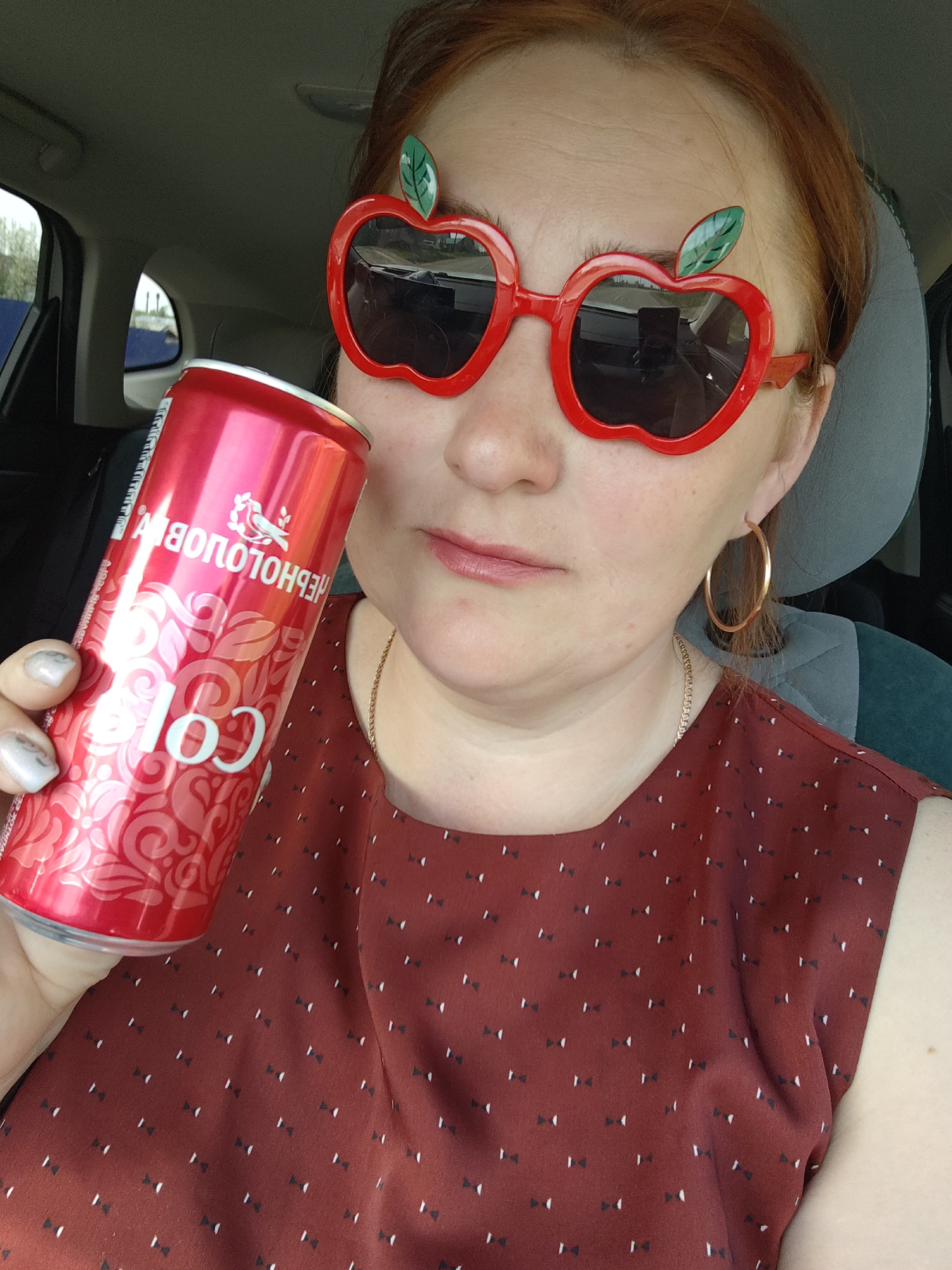 A woman with red hair is indoors, possibly in a car, holding a red can of "Chernogolovka Cola" with a decorative floral pattern. She's wearing sunglasses shaped like red apples with green leaves and has gold hoop earrings. She's also wearing a dark red top with a small geometric pattern. The interior of the car is visible in the background. The location is Фоки, Russia, based on the brand name on the can.