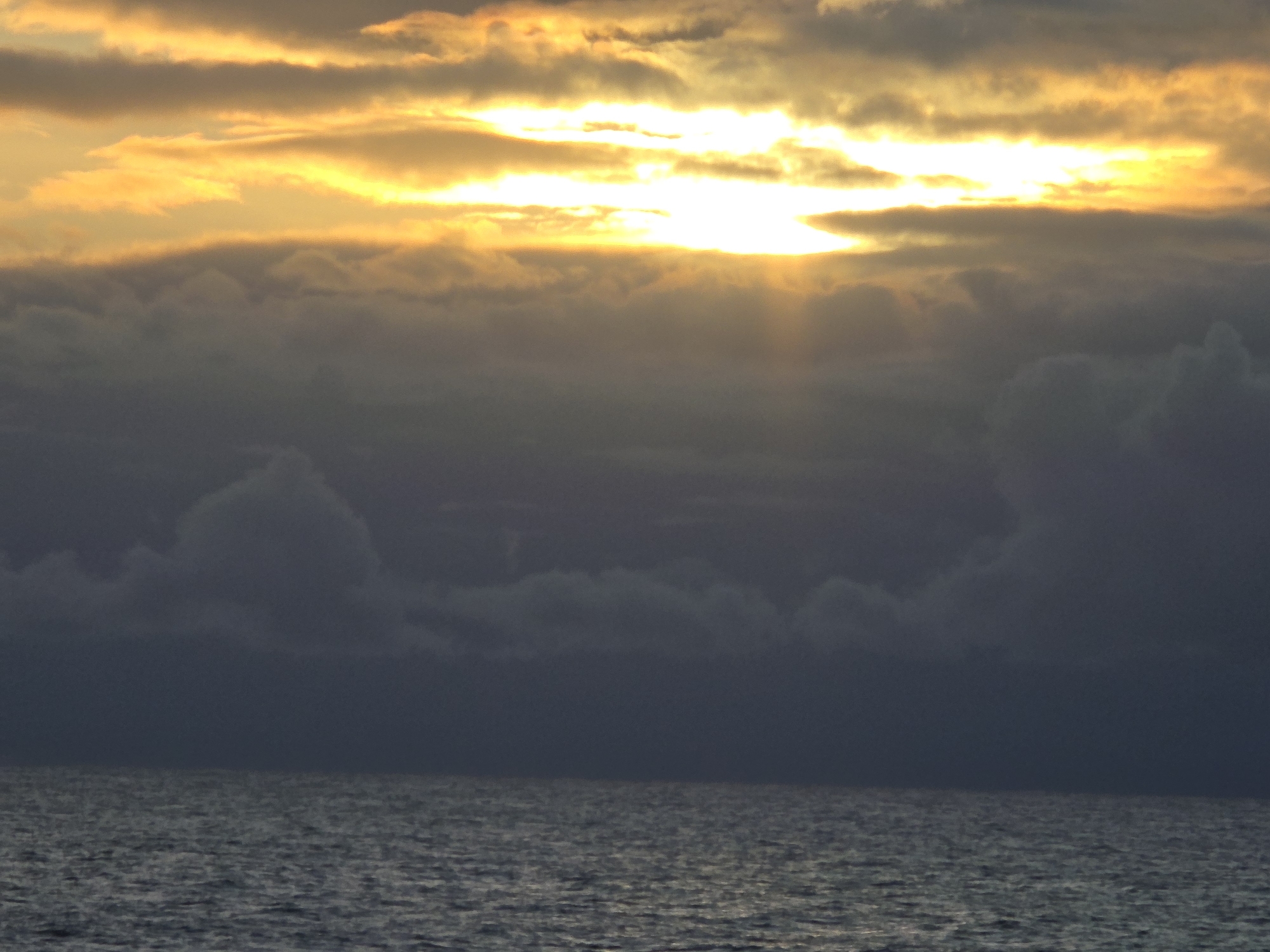 The image depicts a seascape under a cloudy sky. A strip of water occupies the lower third of the frame, displaying a slight rippling effect on the surface. Above the water, a dark band of clouds fills the middle portion, with several puffy, cumulus-like formations visible. The upper part of the image features a partially obscured sun, glowing intensely and casting bright yellow light through gaps in the cloud cover. The overall impression is of a potentially stormy day at sea. No specific location can be determined from the image.