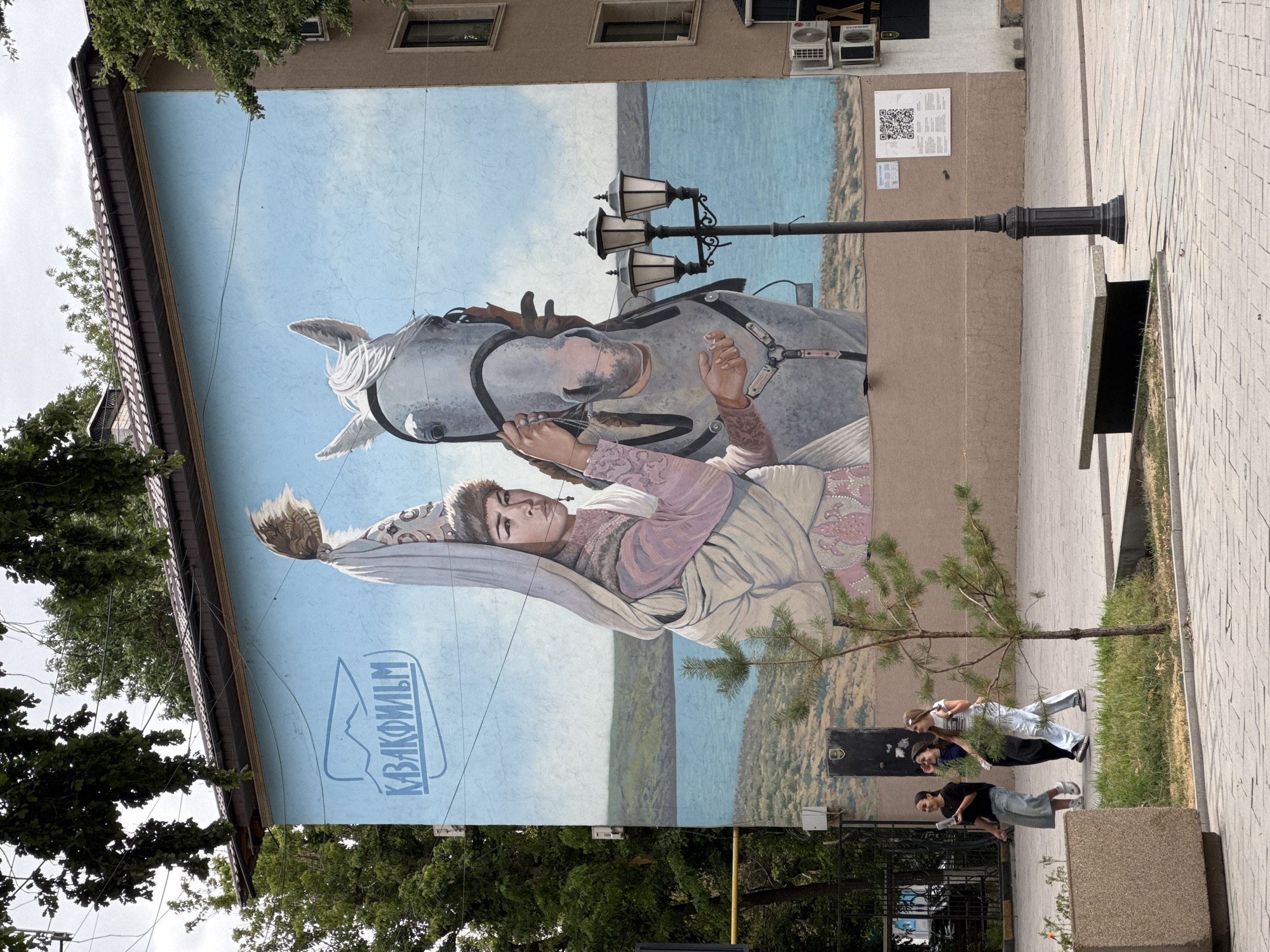 A large mural is painted on the side of a building in the city of Shymkent, Kazakhstan. The mural depicts a woman in traditional attire and a horse, with a lake and hills in the background. The logo "ҚАЗАҚФИЛЬМ" is visible in the upper left corner of the mural. In front of the building, three young women are walking on a paved sidewalk. A lamppost and a bench are also present on the sidewalk. The environment is outdoors, with trees and a cloudy sky.
