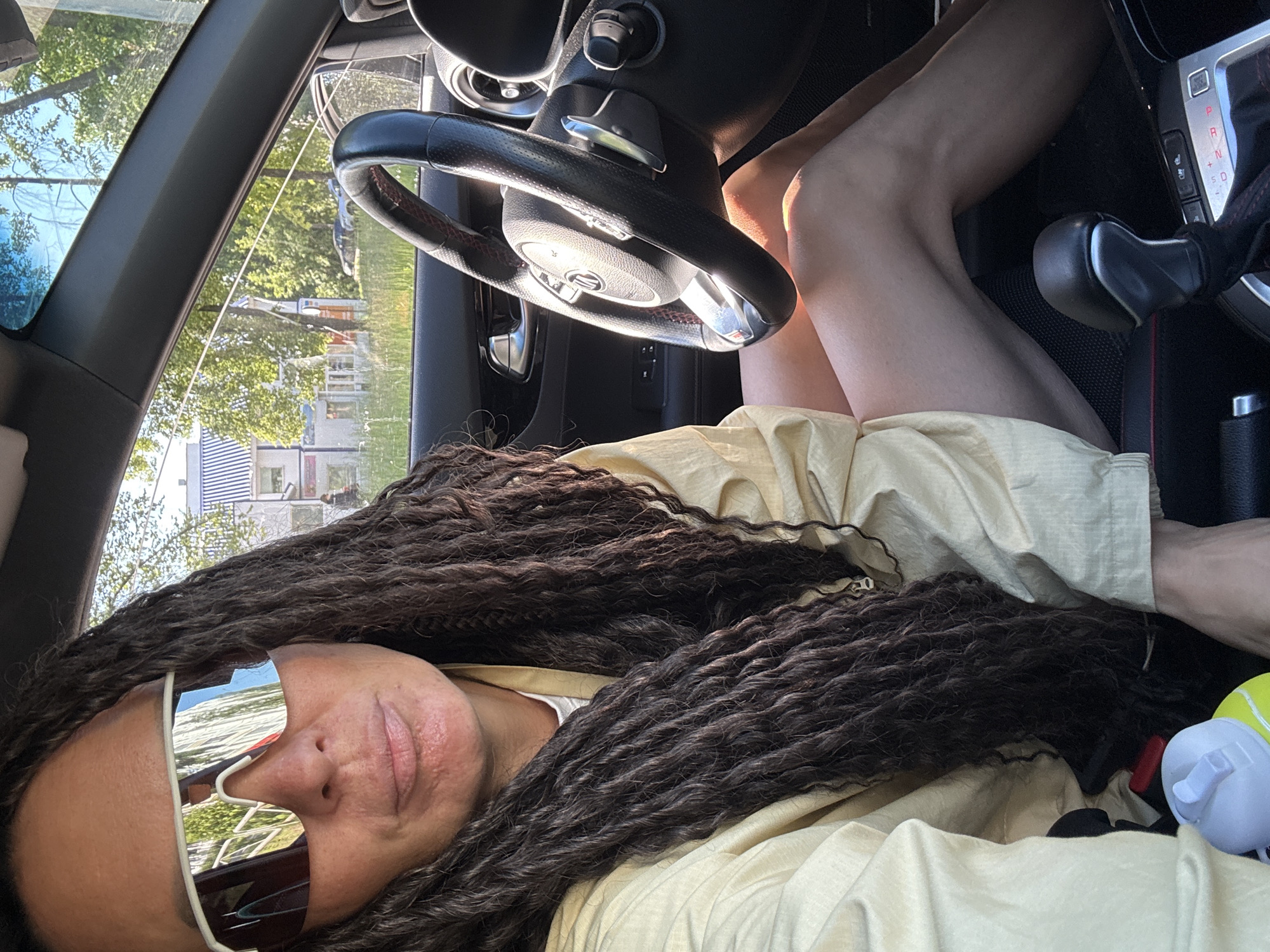 A woman with long, dark, braided hair is sitting in the driver's seat of a car, looking towards the camera. She is wearing large, reflective white sunglasses and a light-colored shirt. Sunlight streams in through the windows, illuminating her legs and the interior of the car. The steering wheel is visible in the foreground, and the gear shift is to the right. Through the windshield, a street scene can be seen, with trees, buildings, and parked cars. The image appears to be taken on a sunny day in Chaikovsky, Russia.