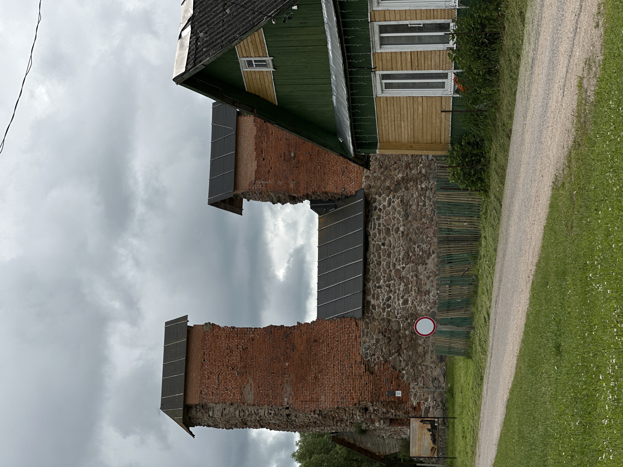 The image shows the remains of a brick tower and a partially intact stone wall, possibly part of a historical ruin or fortification. Above the ruins, a section of a building with a green wooden roof and yellow siding is visible, featuring windows and a small attic window. The building appears to be modern and contrasts with the weathered look of the ruins.
A gravel path runs diagonally across the right side of the image, bordered by a well-maintained green lawn with scattered small white flowers. A dark green wooden fence runs along the stone wall, separating it from the path and lawn.
The sky is overcast with heavy, dark clouds, suggesting a potentially stormy or dull weather condition. A single black wire is visible in the upper left corner of the frame, extending out of view. A circular sign with a red border and a white interior, likely a traffic or informational sign, is mounted on the stone wall.
Below the stone wall, to the left, a wooden information board is partially visible, with what appears to be a map or historical depiction. The overall scene suggests an outdoor location, possibly a park or historical site, under a cloudy sky. There is no clear indication of people or specific activities taking place, other than the presence of a path suggesting pedestrian access. The combination of historical ruins and a more modern building creates a layered historical and visual context.