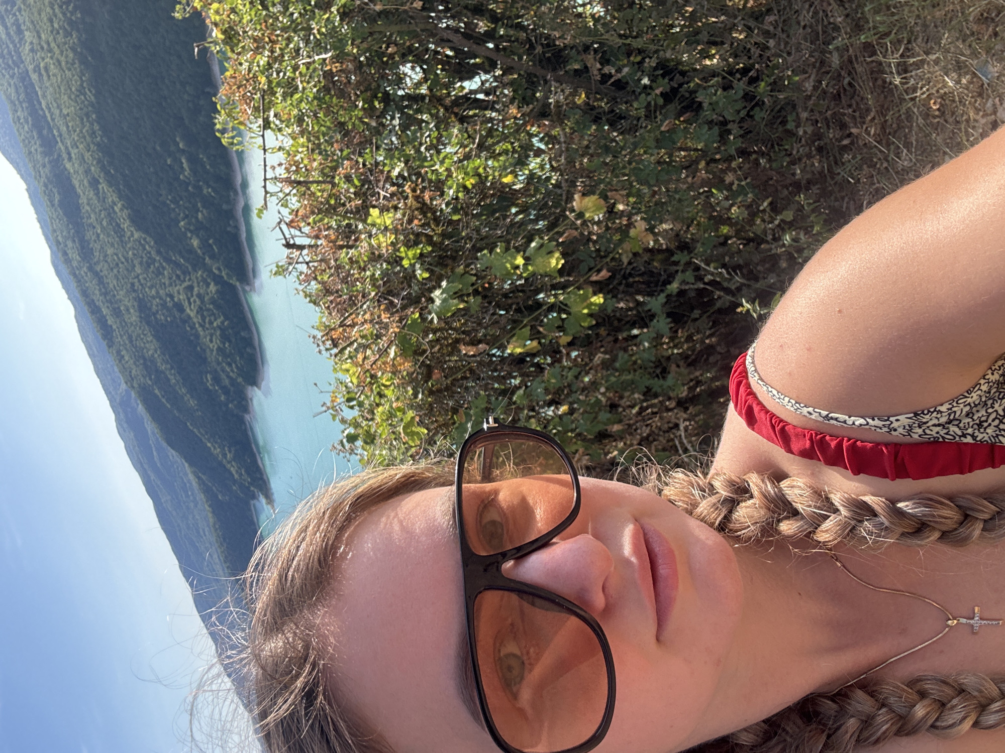 A young woman with blonde hair in two braids and wearing large, dark-rimmed sunglasses with orange lenses takes a selfie. She is wearing a red top with a patterned strap. A gold necklace with a cross pendant is visible. Behind her, a bright turquoise lake is nestled between tree-covered mountains under a clear blue sky. A dense hedge of green and brown foliage is in the foreground, obscuring some of the view. The image appears to have been taken on a sunny day. The location is Moirans-en-Montagne, France.