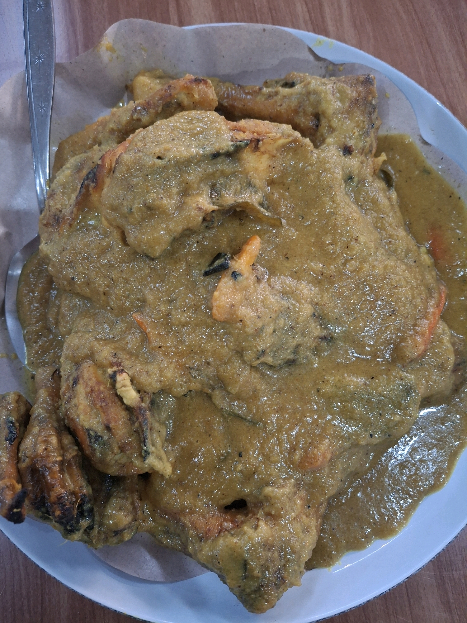 A close-up, overhead shot captures a plate of food served on brown paper, placed on a wooden table. The dish appears to be chicken or seafood in a thick, yellowish-brown curry sauce. Several pieces of the main ingredient, coated in the sauce, are visible. A silver fork rests on the left side of the plate. The lighting suggests an indoor setting. This dish is likely served at a local eatery in Purworejo, Indonesia.