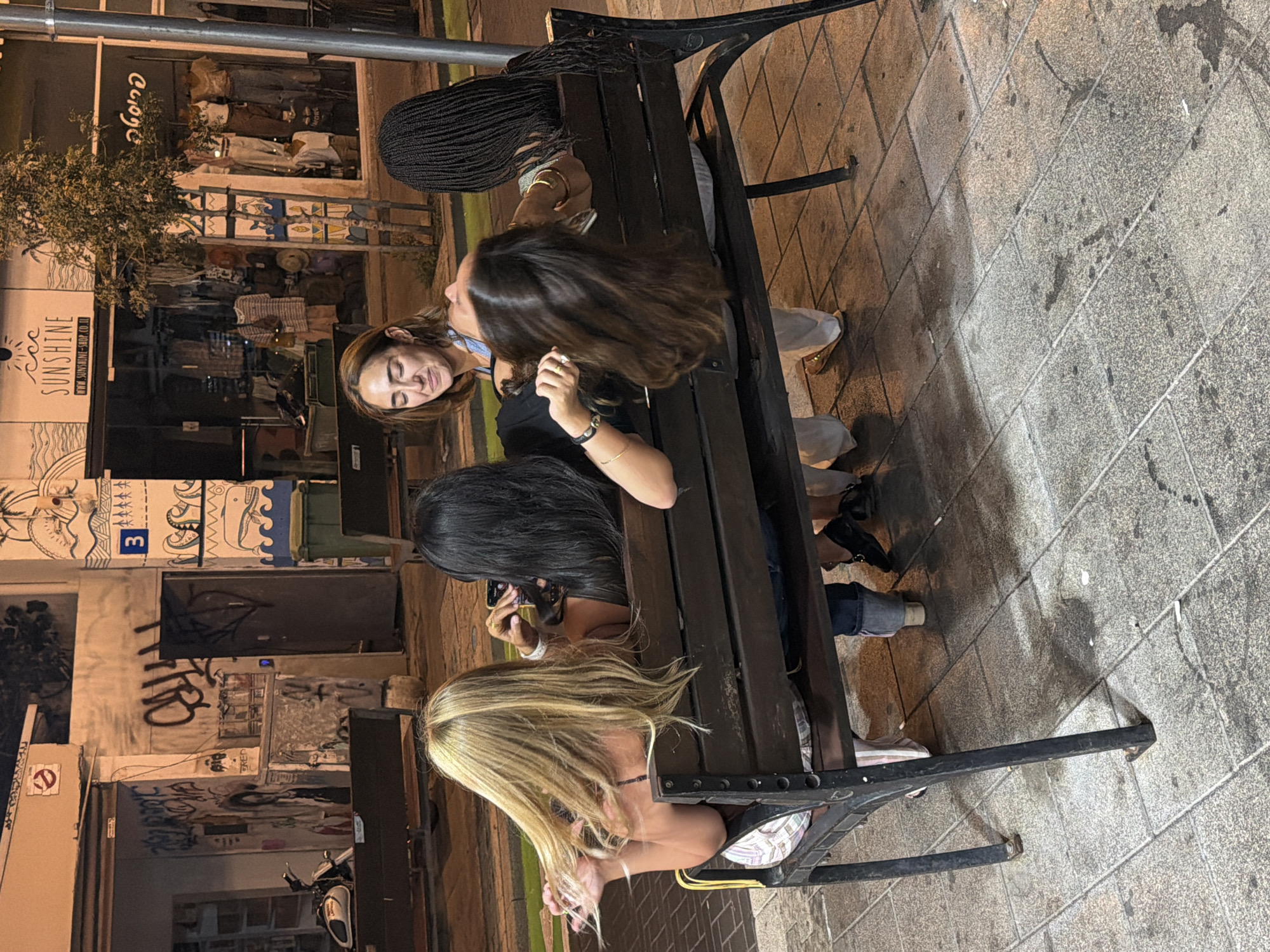 The image depicts four individuals, three women and one person whose gender is not clearly discernible, seated on a dark wooden bench in what appears to be an urban setting at night. The bench is positioned on a tiled sidewalk, with a street visible in the background.
The woman on the far left has long, blonde hair and is wearing a light-colored patterned skirt and a dark top. She is looking to her left.
The second person from the left has long, dark hair and is wearing a dark top. She is holding a smartphone in her right hand and appears to be taking a photo or video.
The third person from the left is facing away from the camera, with her head tilted back. She has long, dark brown hair and is wearing a light blue top and white pants. She has jewelry on her wrist.
The person on the far right has dark, braided hair and is wearing a dark top. They are looking to their left.
In the background, there is a building with a storefront displaying clothing. A sign above the store reads "SUNSHINE" with a website address below. There is graffiti on a wall to the left of the storefront. The scene suggests a public space in Tel-Aviv, Israel, possibly an area with shops and cafes. The lighting is dim, indicating it is evening or night. The overall atmosphere appears casual and relaxed.