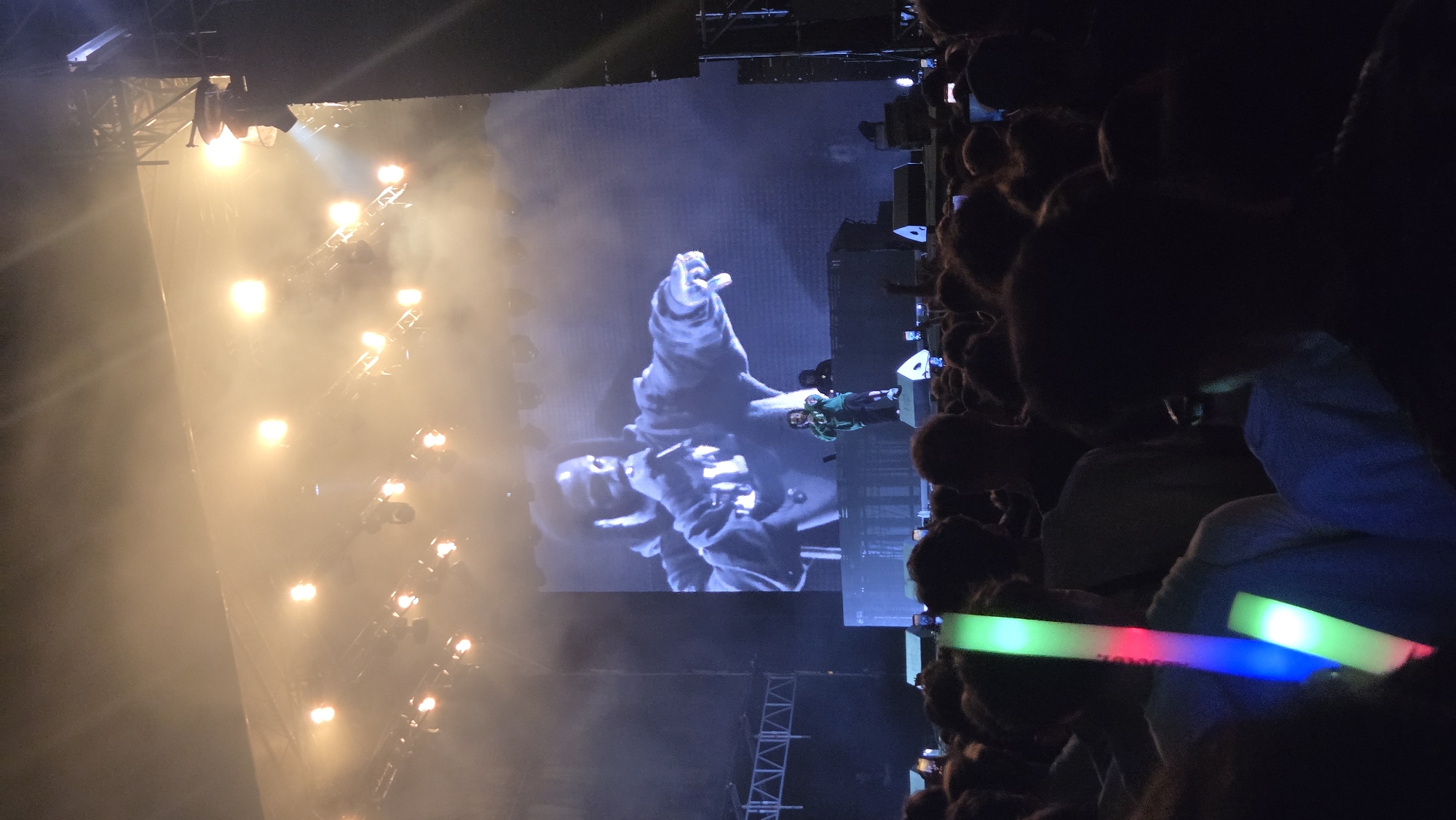 The image shows a concert in Cluj-Napoca, Romania, at the Untold Festival. In the background, a large screen displays a black and white image of a male performer, likely the headliner, holding a microphone. On the stage below, another performer in a green jacket is visible, interacting with the audience. The stage is lit with bright lights and smoke. In the foreground, a crowd of concertgoers can be seen, many holding up glow sticks. The atmosphere appears energetic and festive. The lighting is predominantly stage lights and the glow from the crowd's light sticks. The composition focuses on the performance and the audience's engagement.