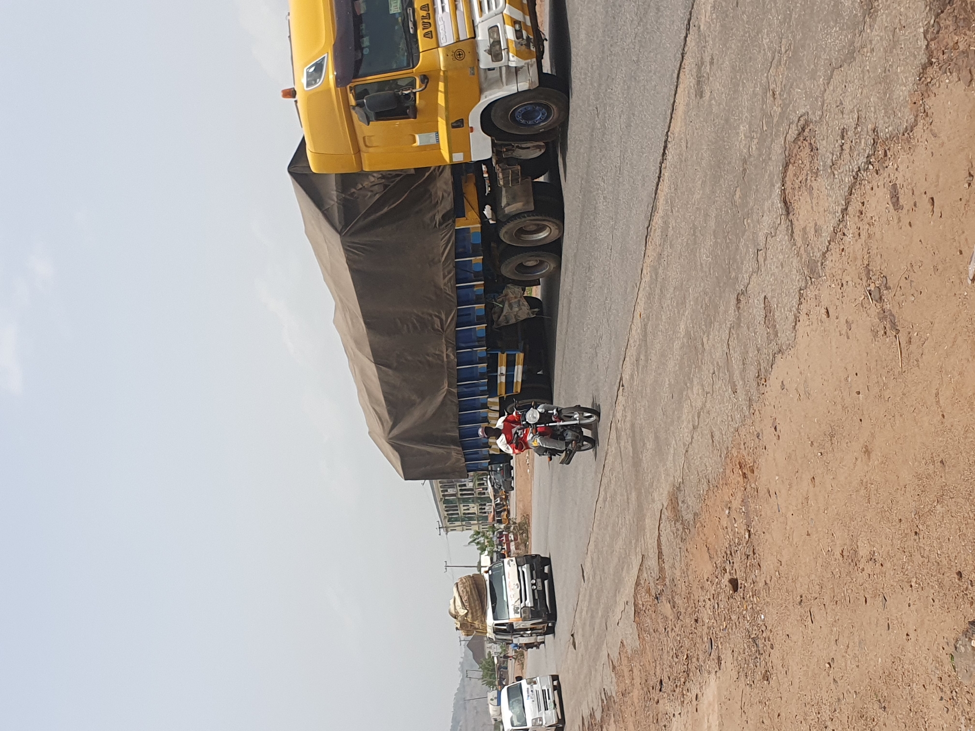 The image captures a daytime road scene in Lokoja, Nigeria, under a bright, hazy sky. A large, yellow semi-truck with a long, dark-tarped trailer is the most prominent feature, positioned on the left side of the paved road. The truck's cab has white and grey accents, and the word "AULA" is partially visible on its side, likely part of a brand name. Below the tarp, blue parts of the trailer's structure can be seen.
To the right of the large truck, a person (or two) on a motorcycle, wearing red and white clothing, is seen traveling on the road. Further down the road, a white pickup truck or van is visible, carrying a large, bundled load on its bed. Another smaller white vehicle appears in the distance, indicating a flow of traffic.
The road surface is paved but shows signs of wear, with a wide, dusty, and uneven dirt shoulder bordering it on the right side. In the background, several buildings, including some multi-story structures with green accents, line the horizon. Distant hills or mountains are faintly visible even further back. The overall scene suggests a busy highway or main road in a semi-urban or rural setting.