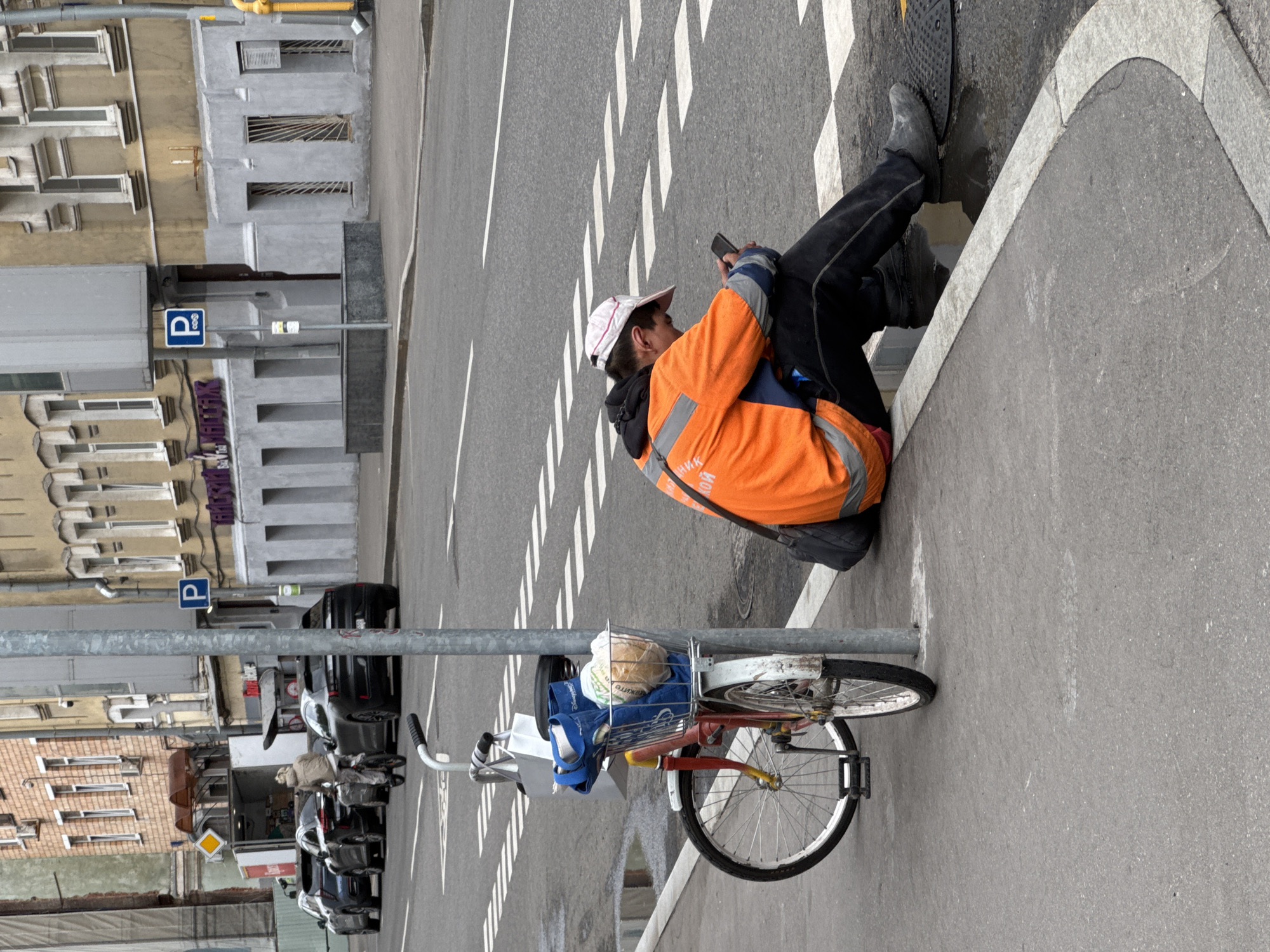 A person wearing an orange high-visibility jacket and dark pants is sitting on a sidewalk next to a parked bicycle, looking at their phone. The person is wearing a light-colored baseball cap. The bicycle has a basket filled with items, including what appears to be bread. The scene takes place on a city street in Moscow, Russia, with cars parked along the side of the road and buildings in the background. A parking sign is visible on a pole. The pavement is wet, suggesting recent rain.