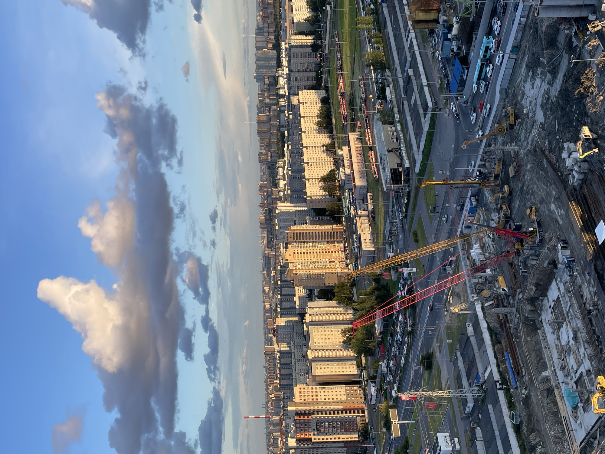 The image shows an aerial view of a city construction site under a cloudy sky. In the foreground, a deep excavation is visible with concrete foundations being laid. Several construction vehicles, including a yellow excavator and two large cranes (one red and one yellow), are present. Further back, a road with traffic, including cars and buses, is seen. Beyond the construction zone, a dense urban landscape unfolds, characterized by numerous apartment buildings of varying heights and architectural styles, some brightly lit by the sun. A prominent red and white striped smokestack is visible in the distance. The sky is a mix of blue with dramatic white and gray clouds, illuminated by what appears to be late afternoon or early evening sunlight. The scene depicts an active construction phase within a developed urban area.