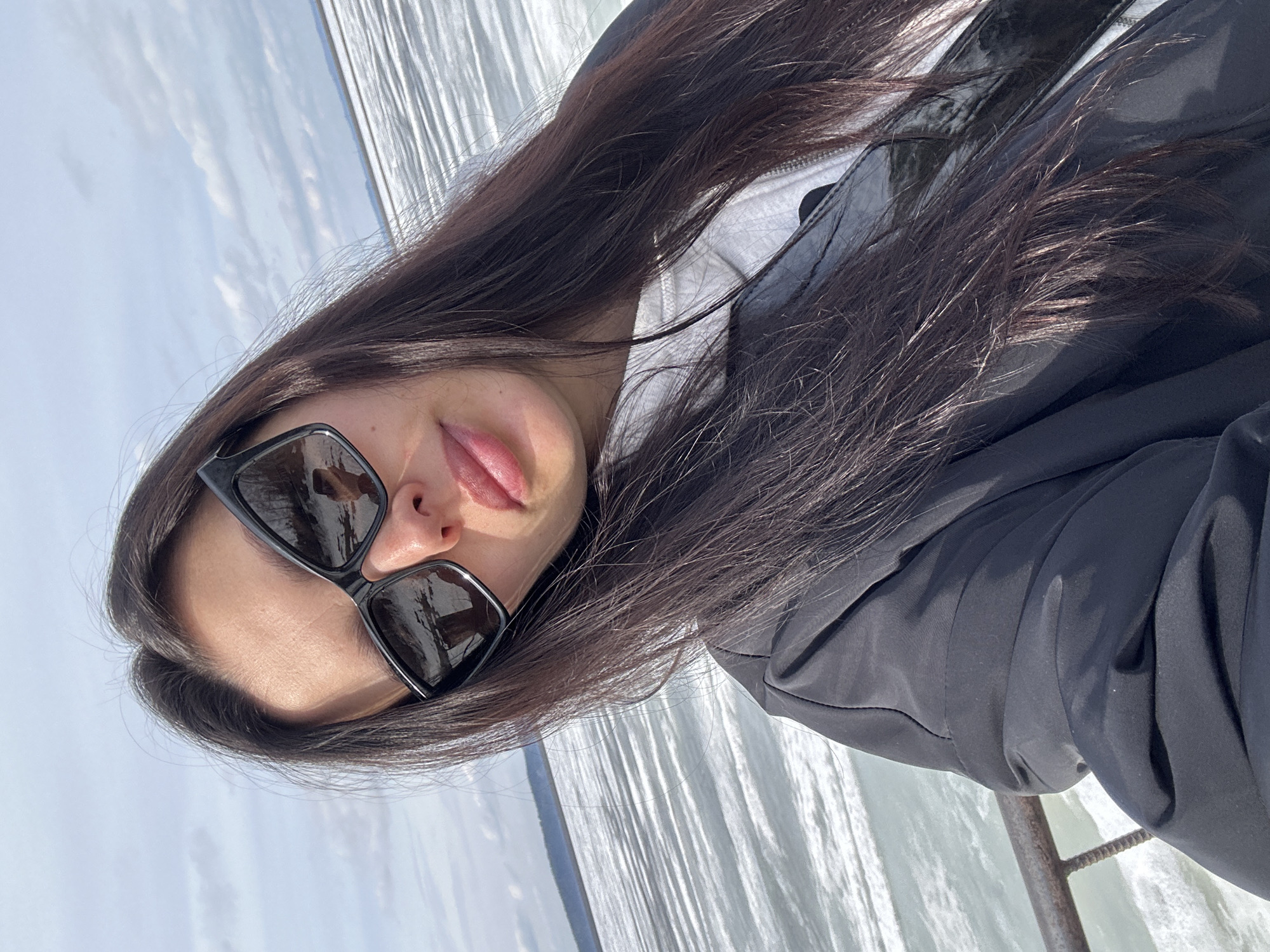 A young woman is captured in a close-up, outdoor portrait, with the image oriented vertically (rotated 90 degrees counter-clockwise). She is positioned on the right side of the frame, looking towards the camera. Her long, dark brown hair flows over her shoulders and chest. She wears dark, rectangular sunglasses that obscure her eyes, and her lips are slightly parted, showing a pink color. She is dressed in a dark, likely black, jacket or coat over a lighter-colored, possibly white or light grey, hooded garment visible at her neck.
The background prominently features a large body of water, which occupies the left and central parts of the image. The water surface shows distinct ripples and reflections, indicating bright daylight. Above the water, in the upper left, a clear blue sky with scattered white clouds completes the outdoor scene. A dark, weathered metal railing or structure is partially visible behind her right shoulder in the lower right corner, suggesting a platform or viewpoint overlooking the water.
The setting is a sunny, daytime environment, consistent with a clear weather day. Given the location context of Chaikovsky, Russia, the expansive body of water is characteristic of the Kama River or the Votkinsk Reservoir. The woman's expression, though partially hidden by sunglasses, appears relaxed. There is no visible text in the image.