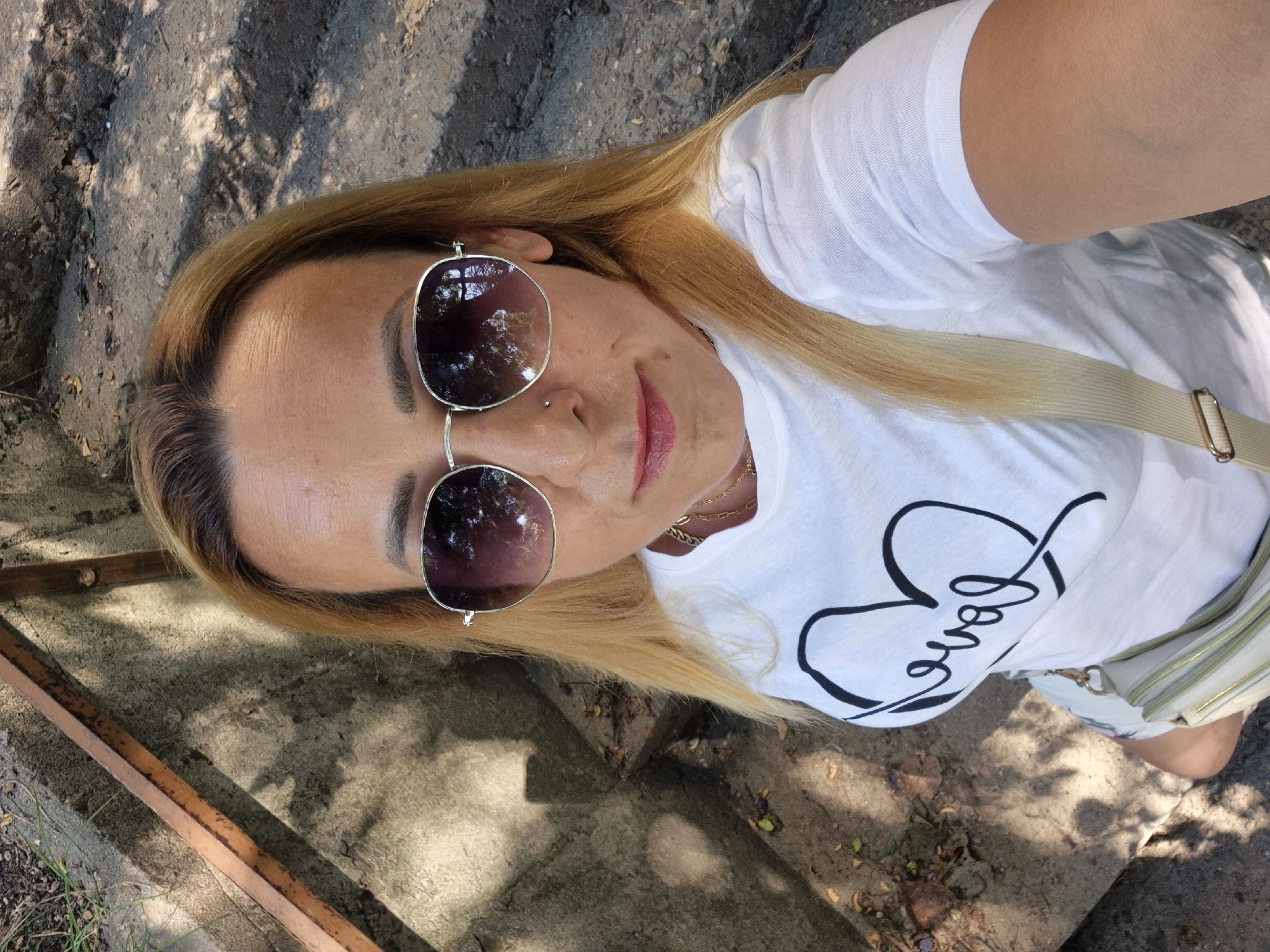 This is a close-up, overhead, selfie-style shot of a blonde woman wearing sunglasses and a white t-shirt with the word "Love" written in a script font within a heart outline. She has on lip gloss and is wearing a gold necklace. A light-colored purse with a gold strap is visible. She is outdoors, as indicated by the shadows on the ground, which appear to be from trees and foliage. The ground itself is a mixture of dirt and concrete. The lighting suggests it is daytime, possibly late morning or afternoon, with the sun casting dappled shadows. The overall mood seems relaxed and casual.