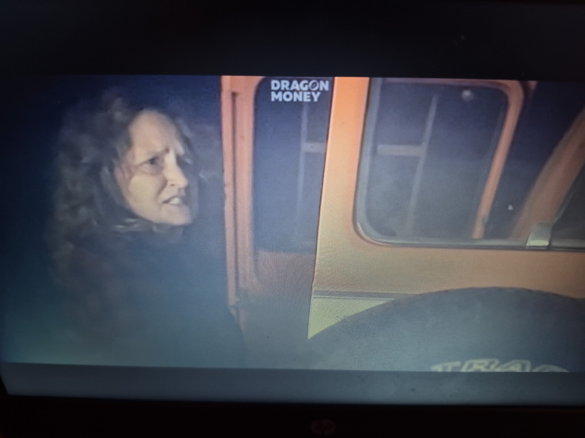 The image shows a still frame from a video, likely a television program, with the words "DRAGON MONEY" visible in white text on a black background in the upper right portion of the frame. To the left of the text is the side profile of a woman with curly, dark hair, her face contorted in an expression of distress or shock. She appears to be looking out of the frame to the left. She is wearing dark clothing. To the right of the woman is the interior of what appears to be a vehicle or booth, with orange paneling and dark windows. Part of a tire is visible in the lower right of the frame, with the word "TRA" partially visible on it. The scene appears to be dimly lit. There is no indication of the specific location within Sandwell, United Kingdom.