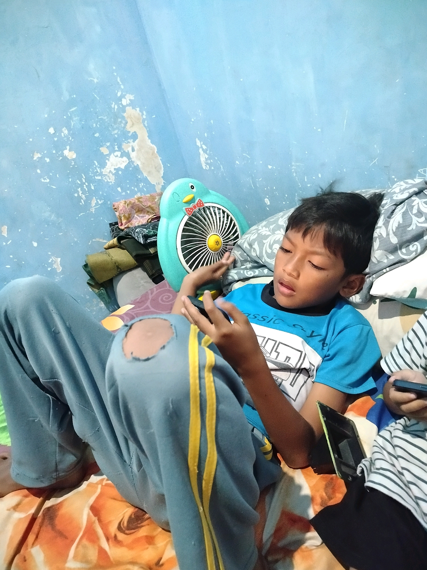 The image shows a young boy sitting on a bed, engrossed in his mobile phone. He is wearing a blue t-shirt and light blue pants with yellow stripes, which are ripped at the knee. The bed is covered with a patterned sheet. Behind him, a turquoise fan shaped like a penguin is visible, along with a stack of clothes and pillows. The wall behind the fan is light blue and shows signs of wear and peeling paint. Another person, possibly a child, is partially visible to the right, also holding a phone. The scene appears to be indoors, likely in a bedroom, in the city of Beloh, Indonesia.