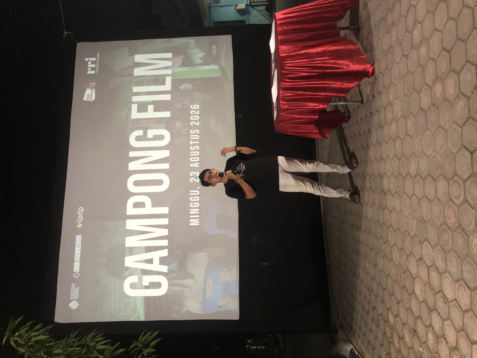 A man stands on a paved courtyard, speaking into a microphone at an outdoor event. He is wearing a black t-shirt and light-colored pants. Behind him, a large screen displays the text "GAMPONG FILM" in large white letters, along with other logos and text, including "KEMENTERIAN KEBUDAYAAN REPUBLIK INDONESIA", "DANA INDONESIANA", "lpdp", "ACEH DOCUMENTER", "rri LHOKSEUMAWE", and "MINGGU, 23 AGUSTUS 2026". A red-draped table with papers on it is positioned to the man's right. The event appears to be taking place at night, as evidenced by the lighting. The courtyard is made of hexagonal paving stones. A plant is visible on the left side of the image, in front of the screen. In the background, a building with a blue roof and a small structure resembling a birdhouse can be seen.