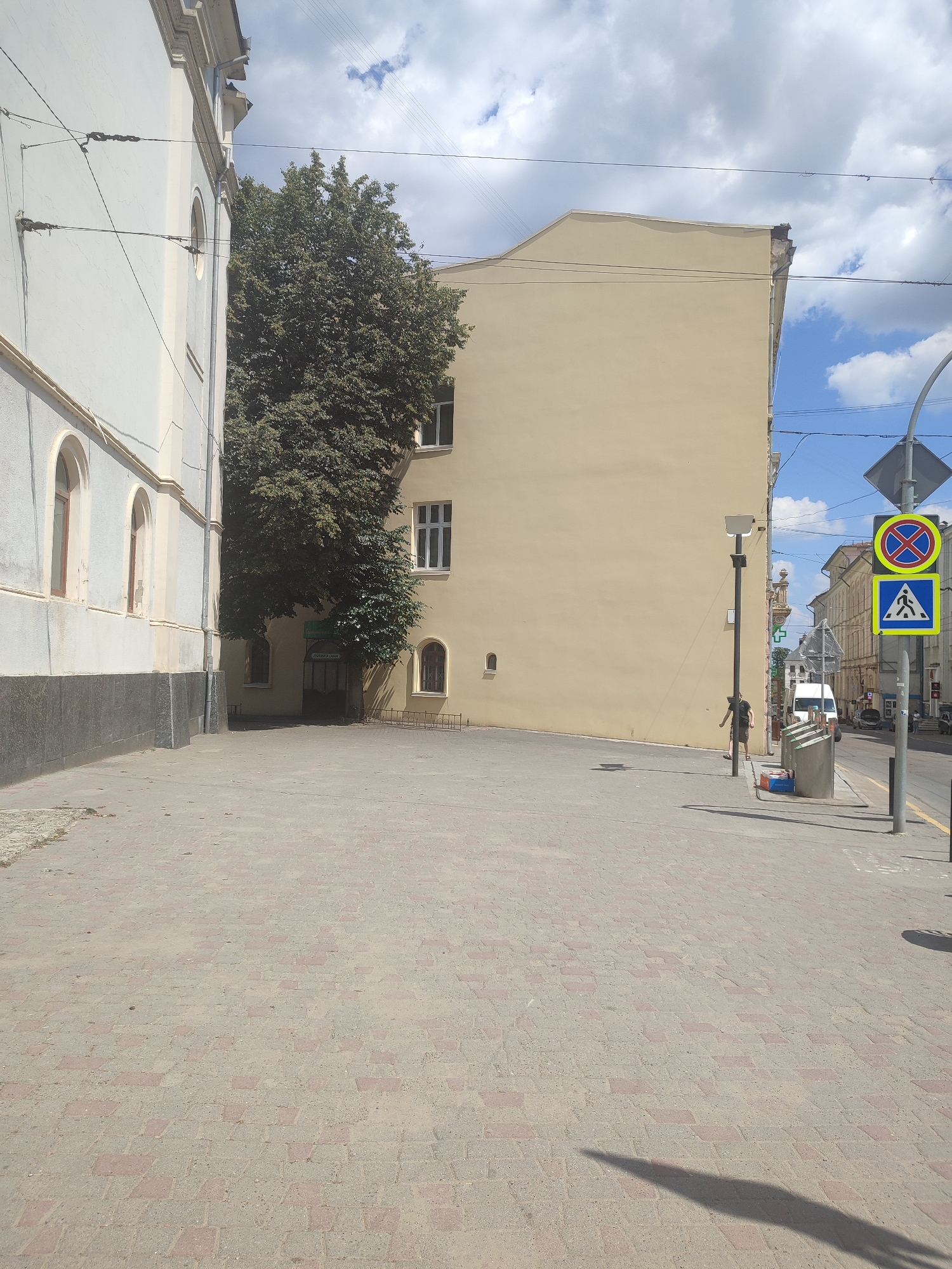The image shows a street scene in Chernivtsi, Ukraine. The foreground is a paved pedestrian walkway with interlocking brick patterns. On the left, a pale blue building with arched windows is visible. To the right of this building, a large tree with dense green foliage stands in front of a tall, light yellow building. The yellow building has several rectangular windows. Further to the right, a street light and a traffic sign pole are prominent. The traffic sign pole displays a circular red and blue sign with a red cross indicating no stopping, and a blue square sign with a white pedestrian symbol indicating a pedestrian crossing. A person in dark clothing is standing near the traffic light pole. Beyond the pedestrian area, a street with vehicles and buildings lining both sides extends into the distance. The sky is partly cloudy with blue patches.