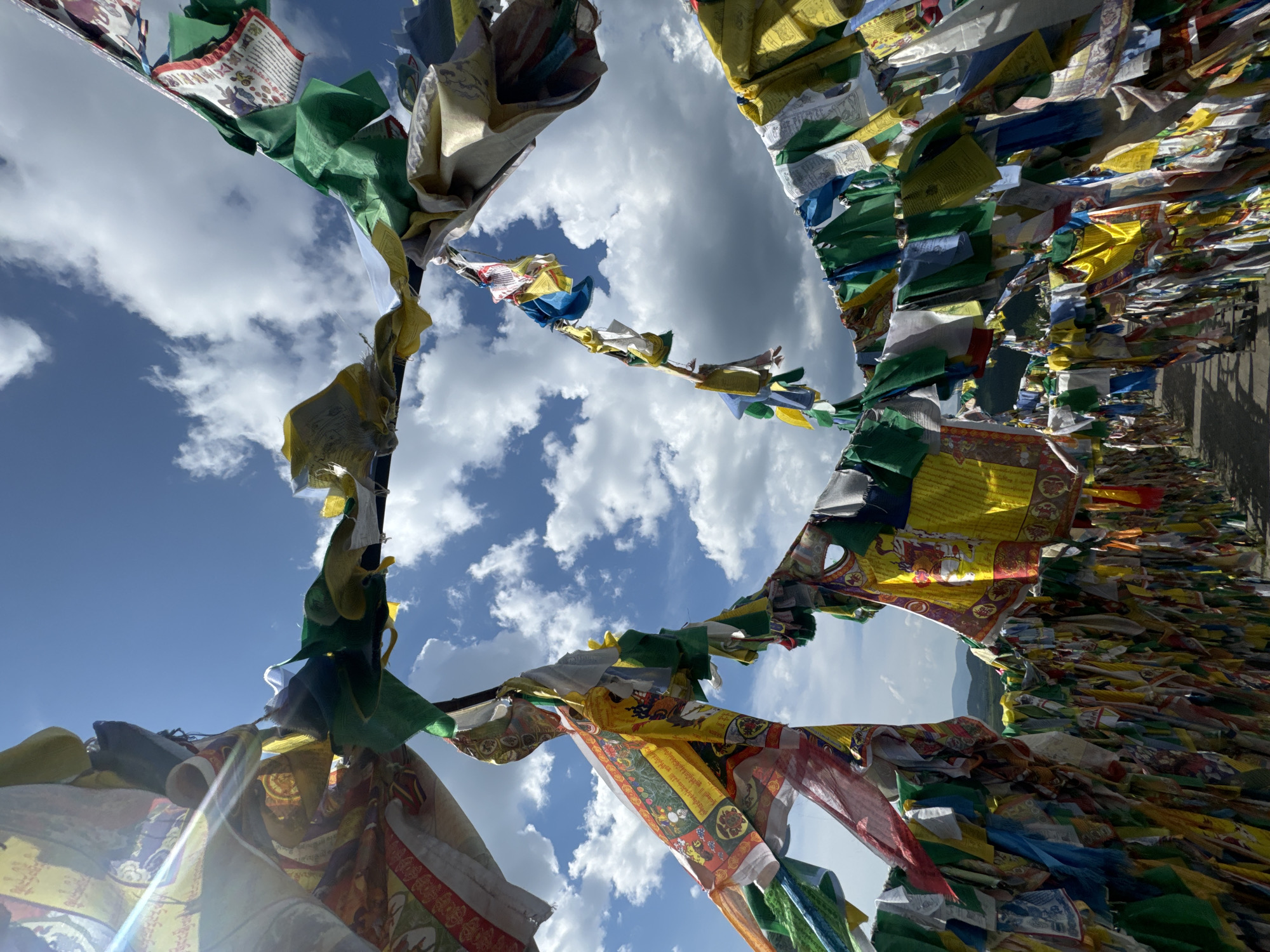 The image shows a path lined with numerous colorful Buddhist prayer flags, fluttering in the wind against a bright blue sky with scattered white clouds. The flags are strung across the path, creating a tunnel-like effect. In the distance, rolling hills are visible under the sky. The scene is located at the Ivolginsky Datsan, a Buddhist monastery located near Ulan-Ude, Russia. The flags are vibrant and varied in color and design, with some featuring intricate patterns and symbols. The sunlight casts shadows on the path, indicating a bright day. There are no people or animals visible in the image.