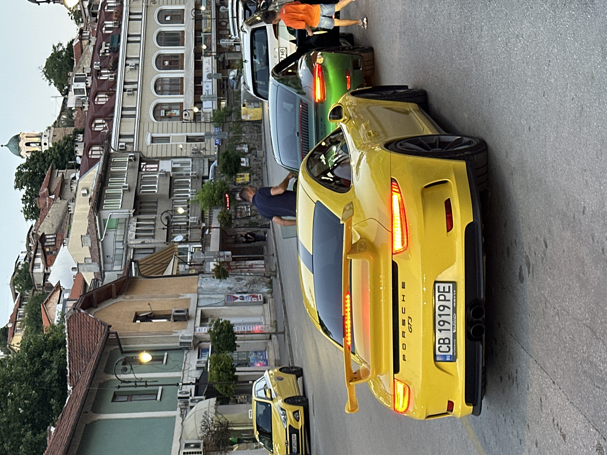The image shows a yellow Porsche GT3 parked on a street in Veliko Tarnovo, Bulgaria. The car is facing away from the camera, and its rear lights are illuminated. A green Porsche is parked behind it. The street is lined with buildings, some of which appear to be old and historic. There are a few people visible in the image, including a man standing near the green Porsche and a child walking away from the camera. A yellow taxi is also visible in the background. The overall scene depicts a street in a European city, likely a tourist destination, with luxury cars present.