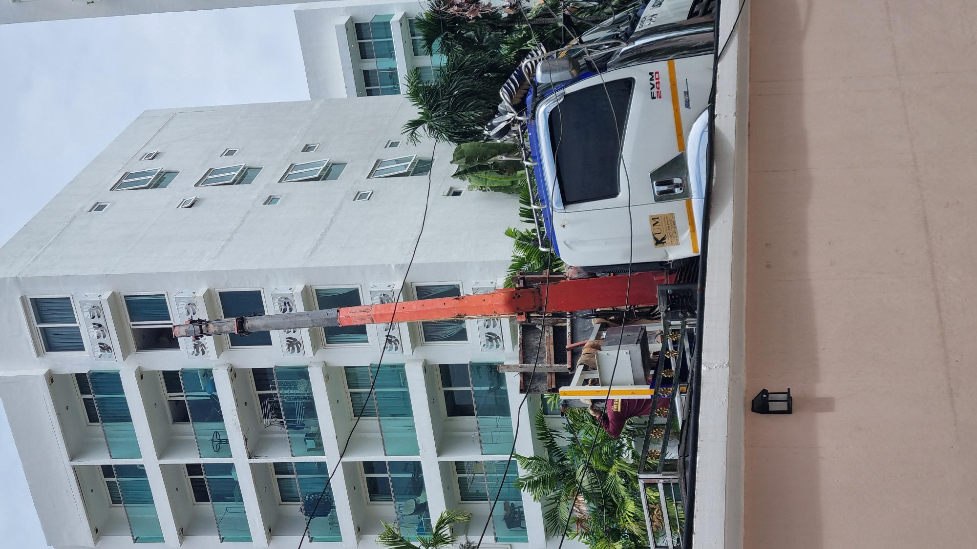 In Pattaya City, Thailand, a crane truck is positioned near a modern white apartment building with multiple balconies and blue glass railings. A worker, wearing a red shirt with text and a tan hat, is visible in the crane's basket, appearing to be working on the side of the building. The truck has a white cab with blue trim and the text "KUM" and "FVM 240" are visible on its side. Lush green palm trees and other foliage surround the base of the building and are partially obscuring the truck. The sky is overcast.