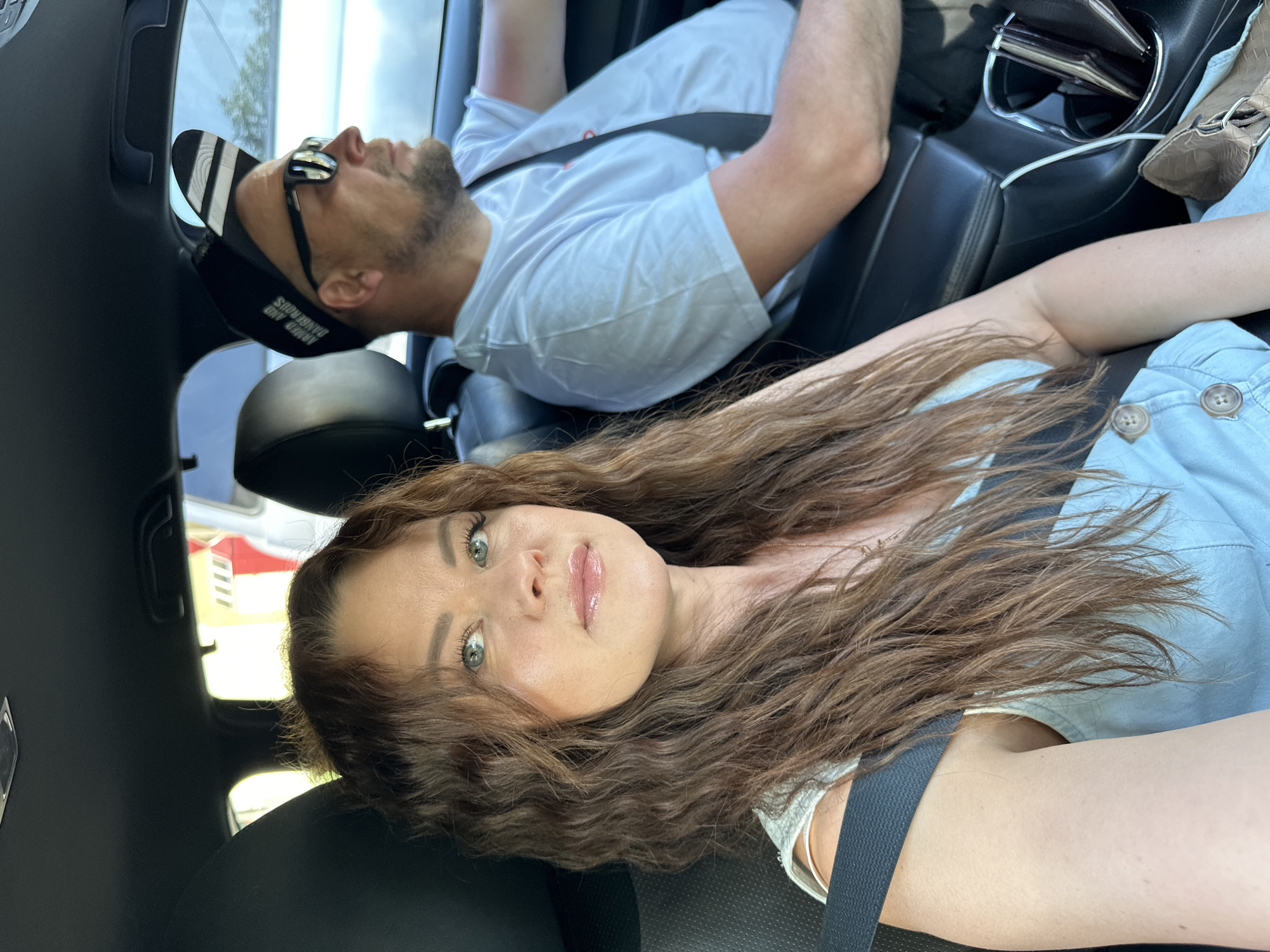 The image shows two people inside a car. The woman is in the foreground, facing the camera with long, wavy brown hair. She's wearing a light blue denim top and a seatbelt. Behind her is a man wearing a white t-shirt, a baseball cap, and sunglasses. He also has a seatbelt on and appears to be reclining in his seat. The interior of the car is visible, showing black leather seats, the center console with a charging cable and a purse. Outside the window is a glimpse of a red and white building, potentially located in the city of Chaikovsky, Russia.