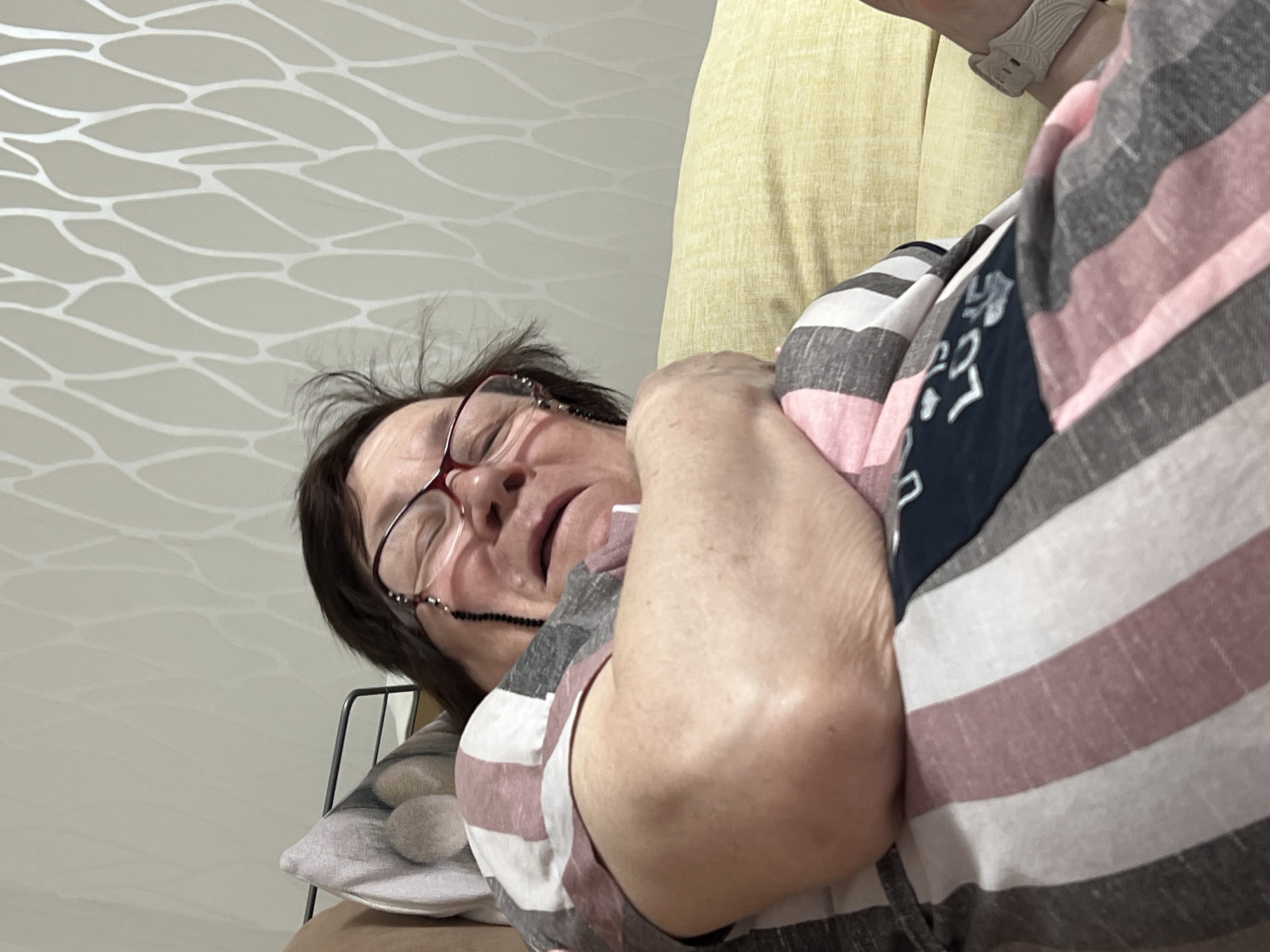 An elderly woman with dark hair, wearing red-framed glasses with a beaded chain, reclines in a chair, possibly asleep or resting. Her mouth is slightly open. She wears a striped shirt with colors including pink, white, and gray. A portion of a navy blue logo is visible on the shirt. A light yellow cushion is behind her. Her left arm is folded across her chest. She has a watch on her wrist. The chair appears to be near a white patterned wall, and a gray cushion with a circular pattern is visible near her head. The location is potentially a residence in Perm, Russia.