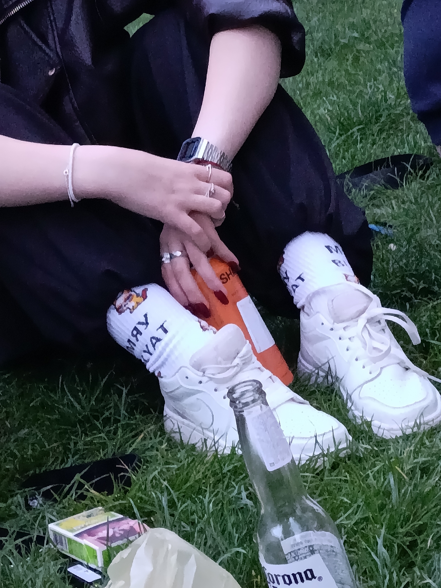 A person sits on grass outdoors. The person wears black pants, white sneakers, and white socks with cartoon graphics and the word "YAYAT". The person wears a watch on their left wrist and rings on both hands. An empty Corona beer bottle and a pack of cigarettes are on the grass near the person's feet. The location is Yerevan, Armenia.